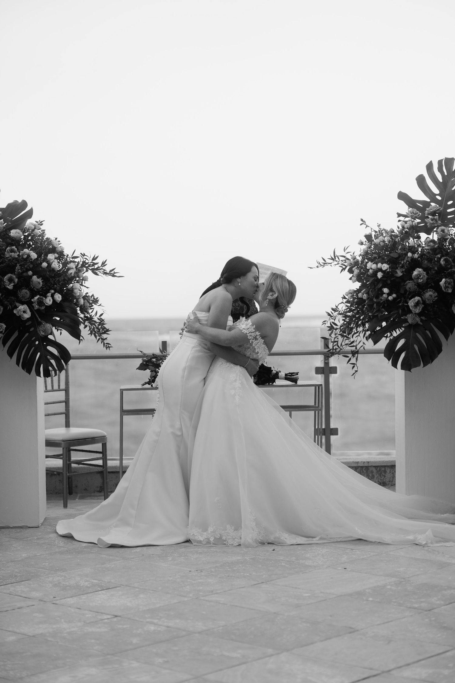 Two brides share a kiss at their outdoor wedding ceremony in a romantic black and white photo.