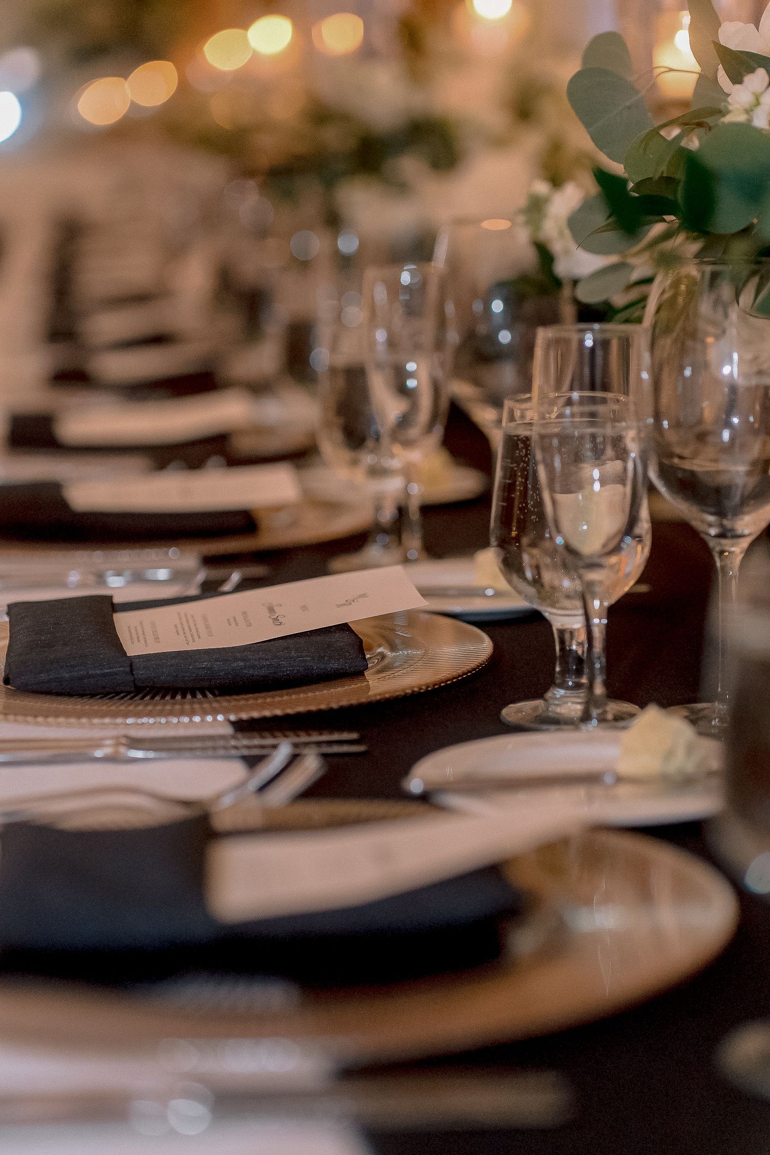 Elegant wedding reception table setting with gold charger plates, black napkins, crystal glasses, and floral centerpieces.