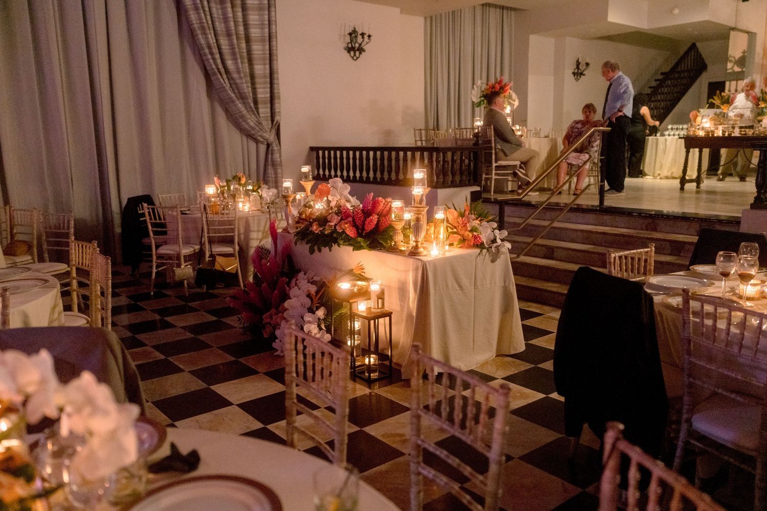 Romantic candlelit wedding reception hall with floral centerpieces on black and white checkered floor.