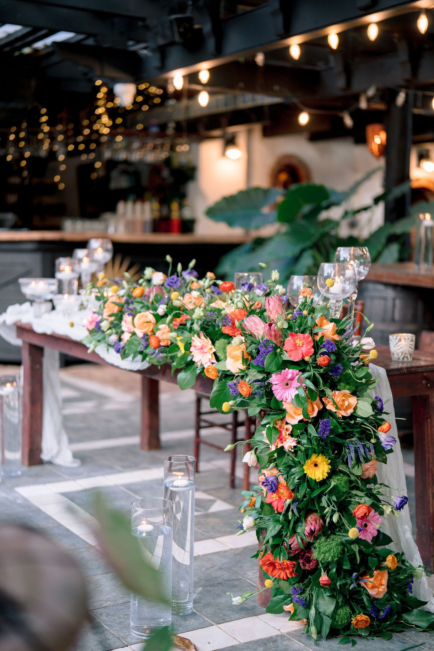 Vibrant floral garland centerpiece with orange, pink, and purple blooms adorning a rustic wooden wedding reception table.