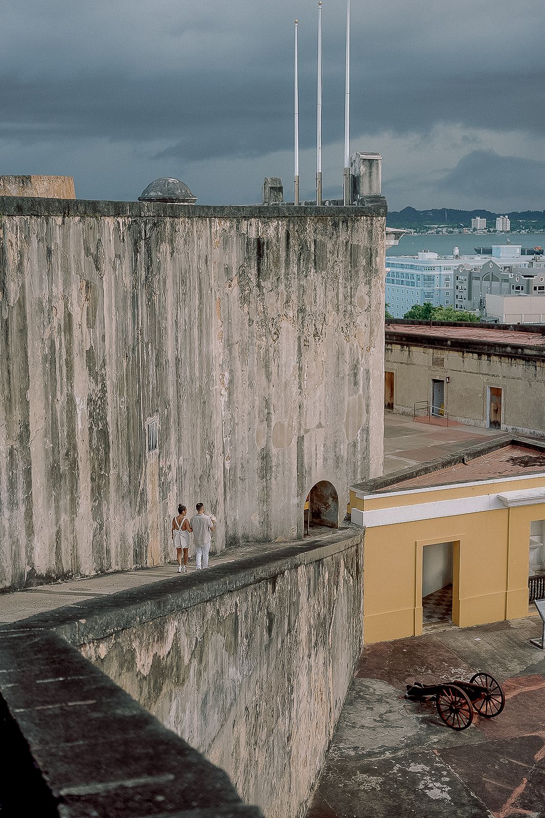 Couple in white standing on ancient fortress wall at Castillo San Cristobal with cannon in courtyard below.