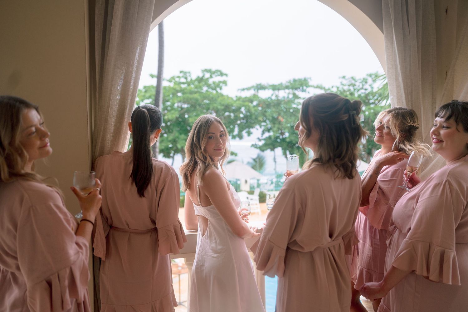 Bride and bridesmaids in pink robes celebrating together near a sunlit window with tropical views.