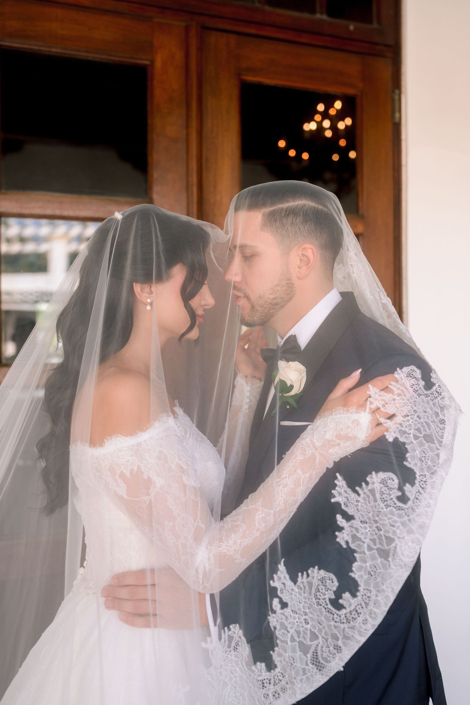 Bride and groom share intimate moment under lace wedding veil, groom in tuxedo, bride in off-shoulder gown.