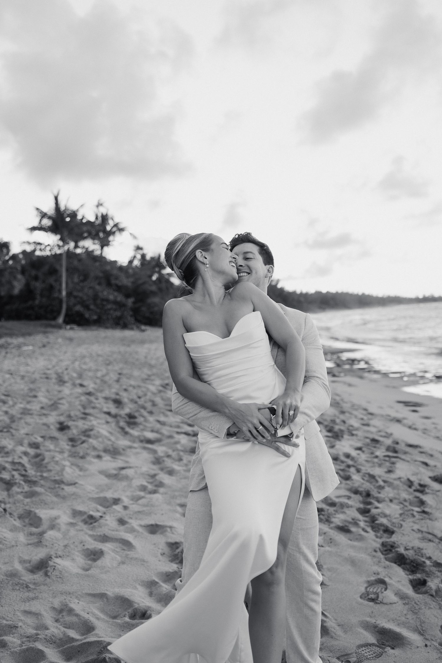 Black and white beach wedding photo of bride and groom embracing on sandy shore with palm trees and ocean in background.