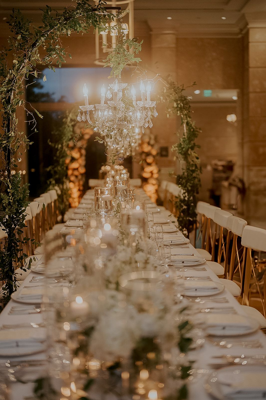 Elegant wedding reception table with crystal chandelier, candles, greenery, and white florals in a warmly lit ballroom.