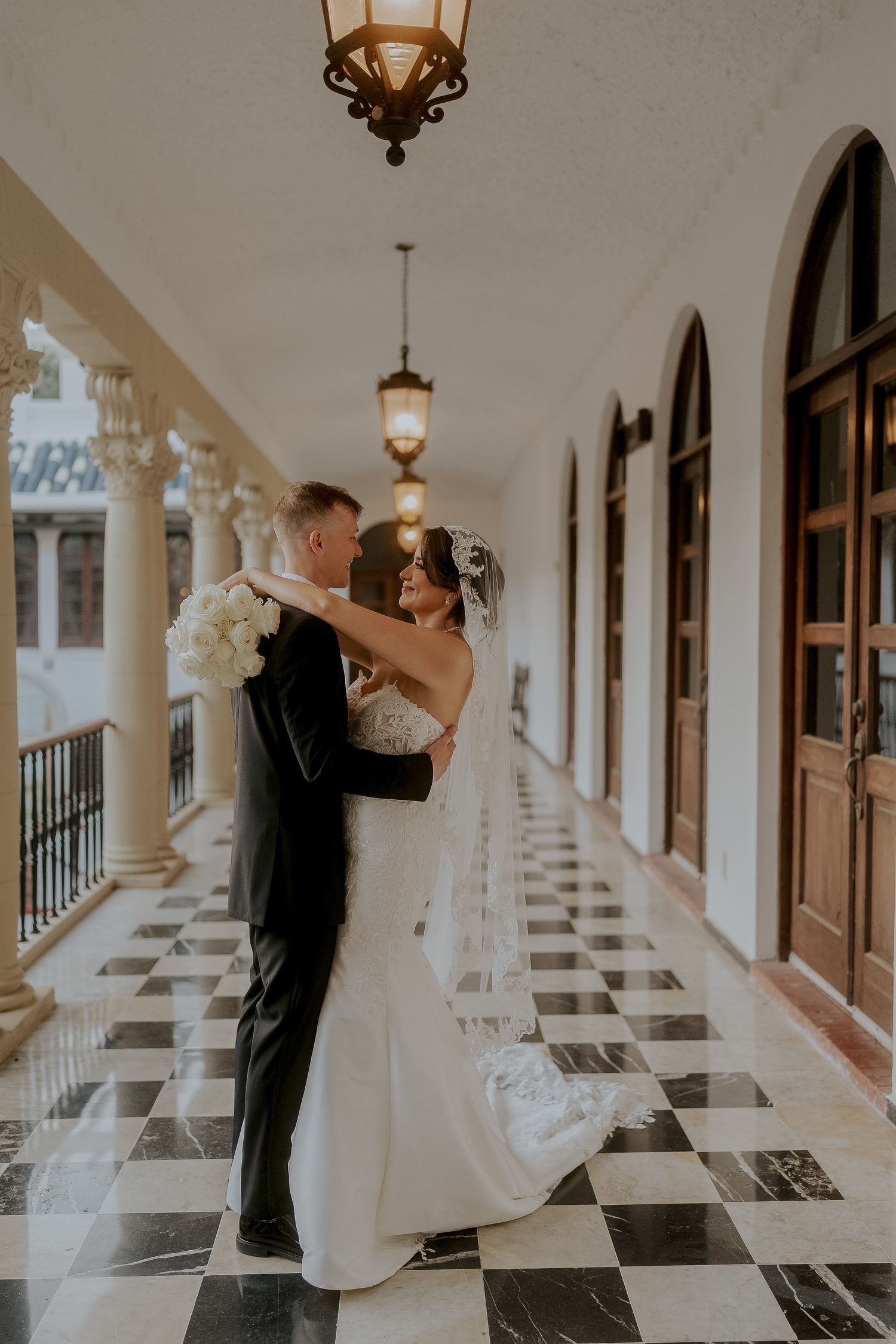 Bride and groom embrace on checkered marble balcony corridor with ornate lanterns at Casa de Espana.