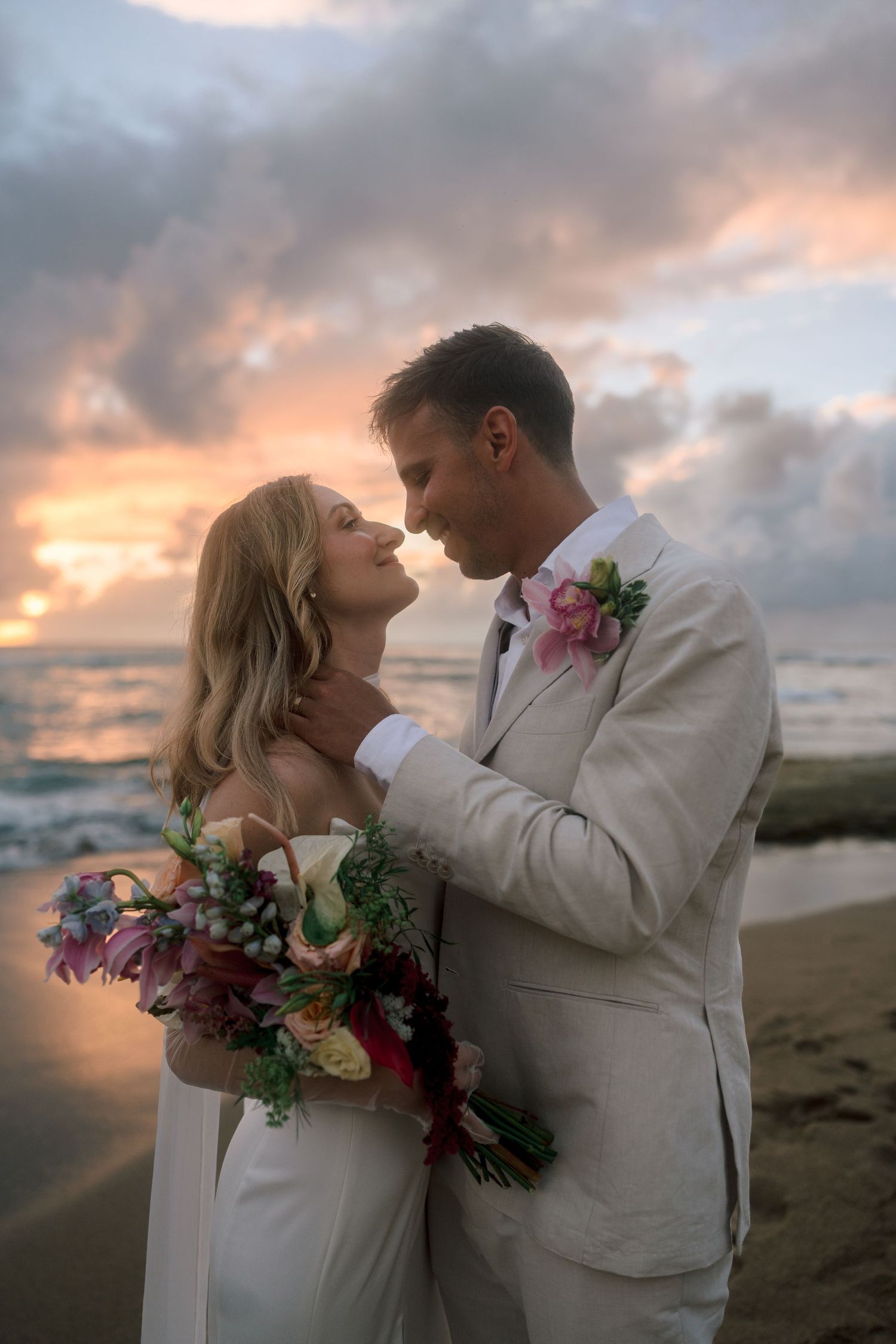 Couple shares a romantic moment on a beach at sunset, bride holding a vibrant tropical bouquet.