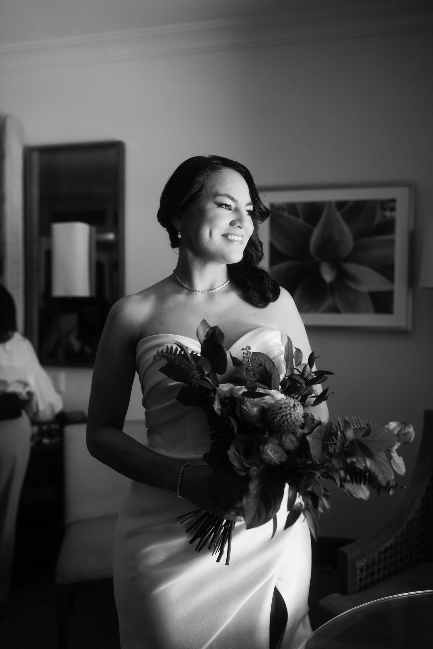 Bride in strapless gown smiling while holding a lush bouquet, captured in elegant black and white photography.