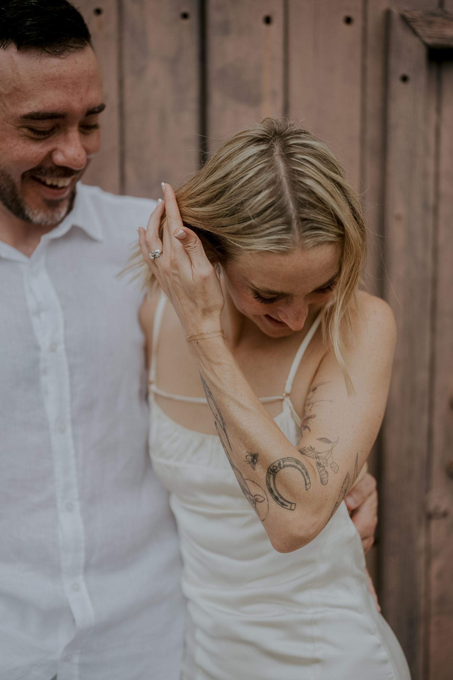 Smiling groom embraces tattooed bride in white dress against rustic wooden wall.