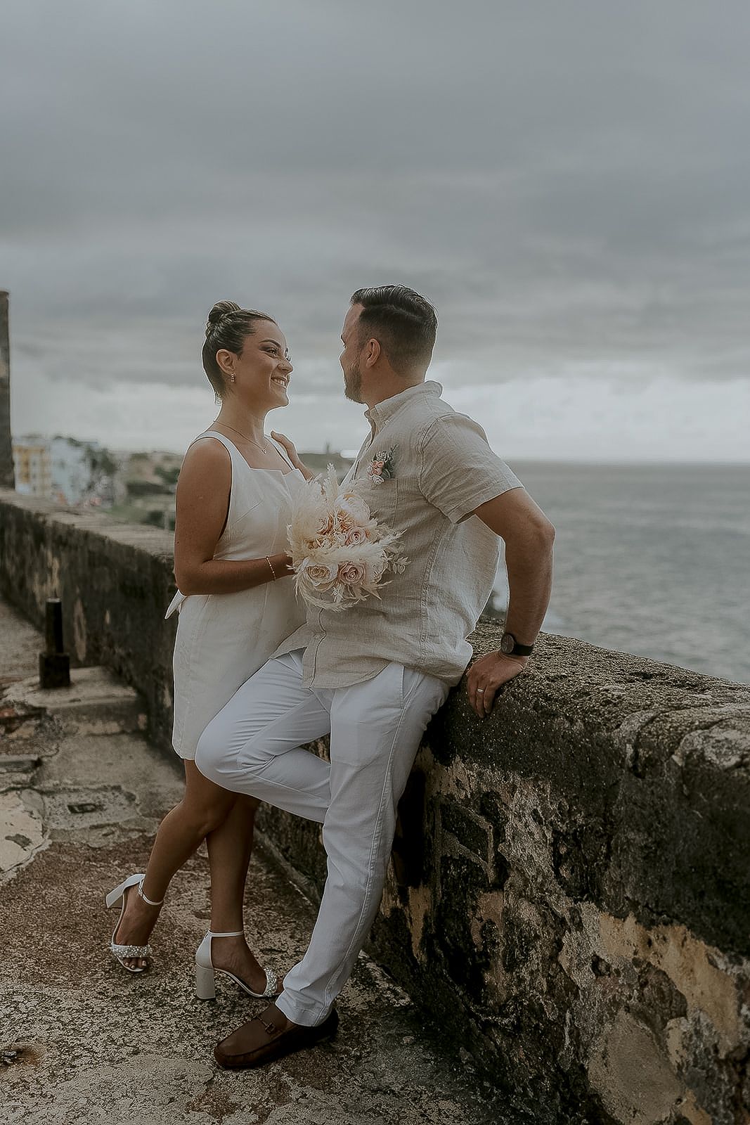 Couple in white wedding attire sharing a romantic moment on a stone wall overlooking the ocean on a cloudy day.