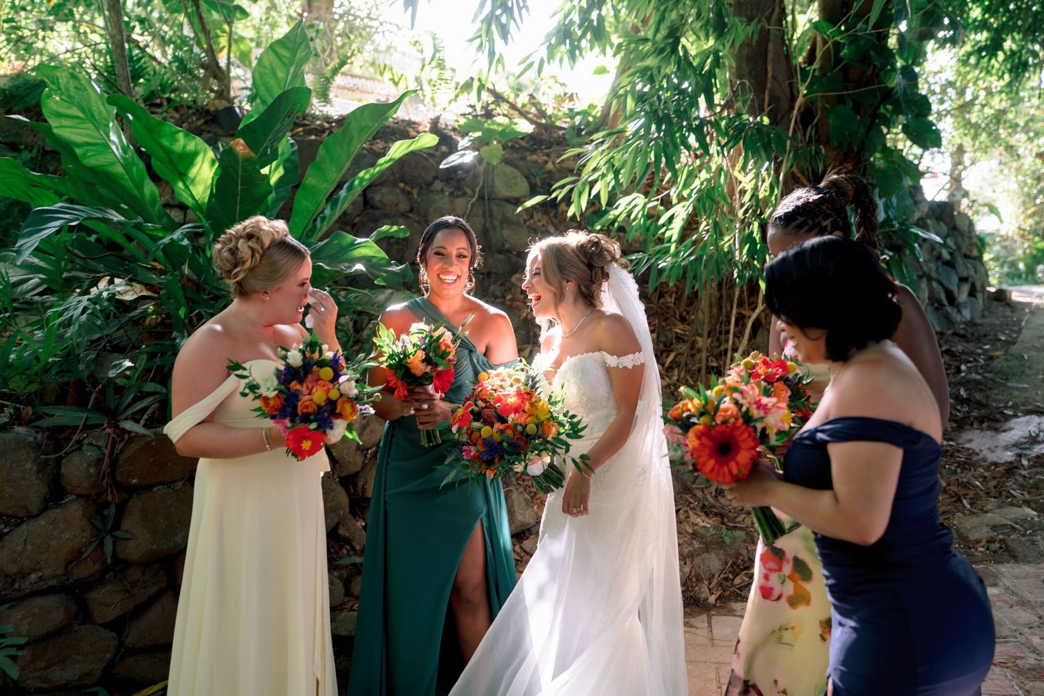 Bride in white gown with bridesmaids in green and navy dresses holding colorful bouquets in a tropical garden setting.