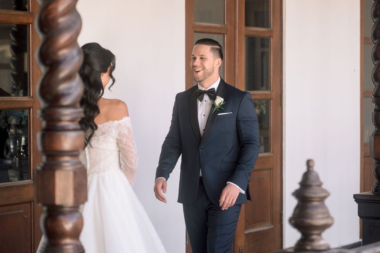 Groom in navy tuxedo smiles at bride in off-shoulder lace wedding dress during first look ceremony.