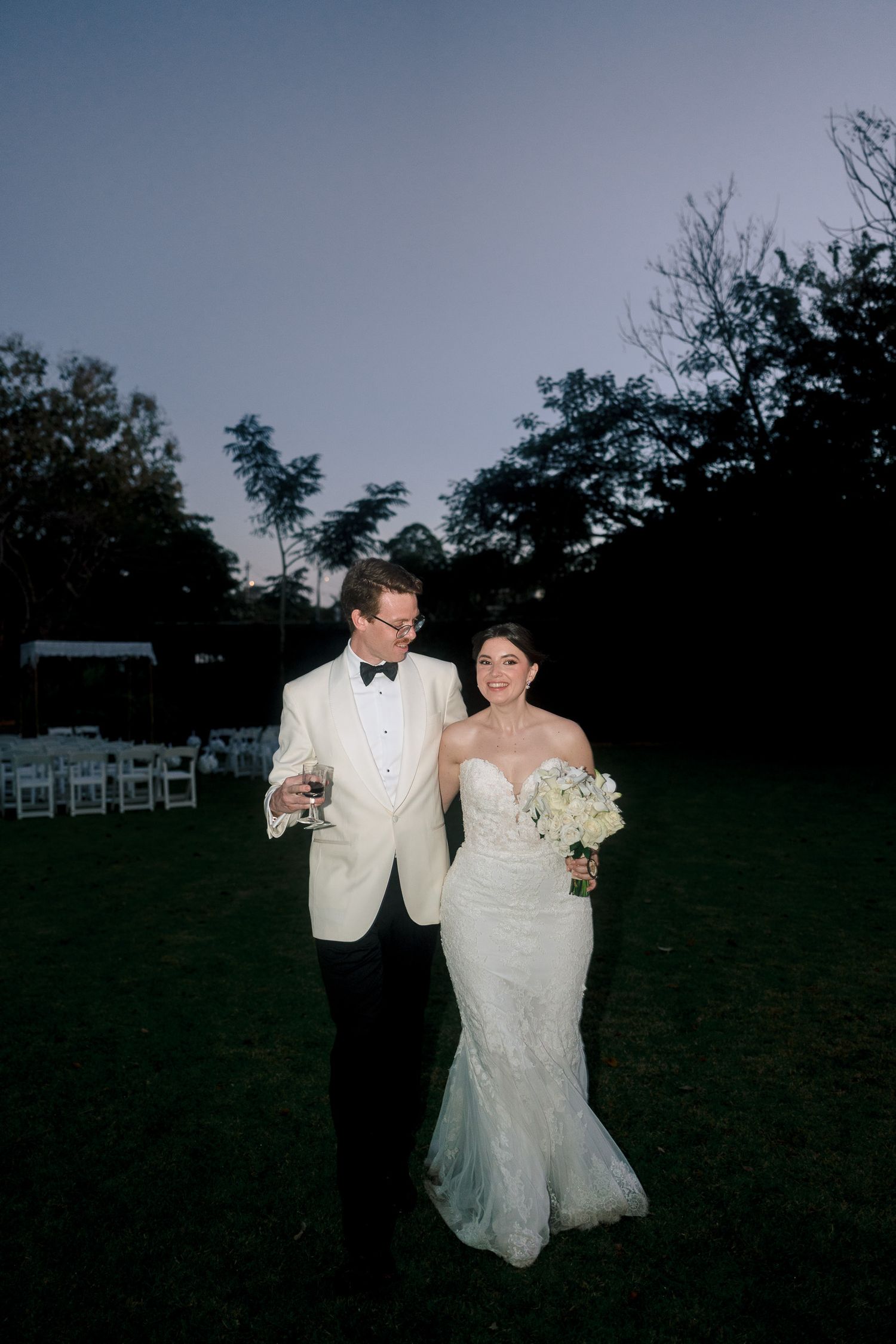 Bride in fitted lace gown and groom in white tuxedo jacket walk together at dusk on lush green lawn.