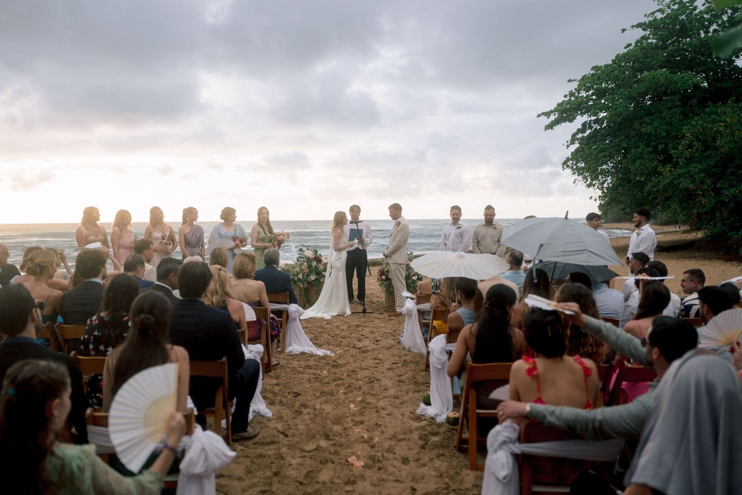 Romantic beach wedding ceremony at sunset with guests seated on sand chairs facing the couple at the altar.