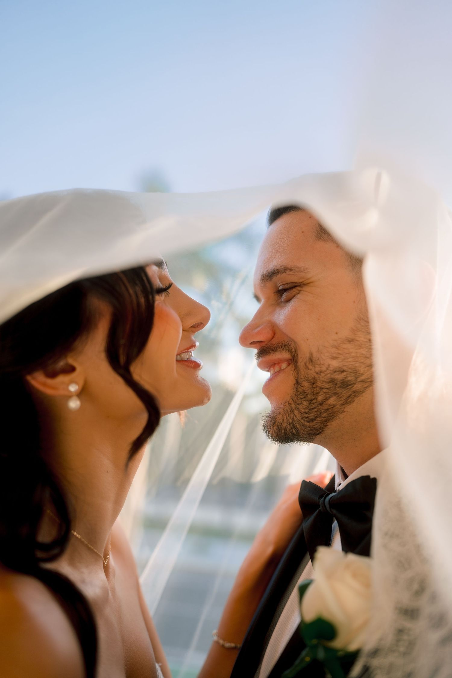 Bride and groom smiling at each other under a white veil on their wedding day.
