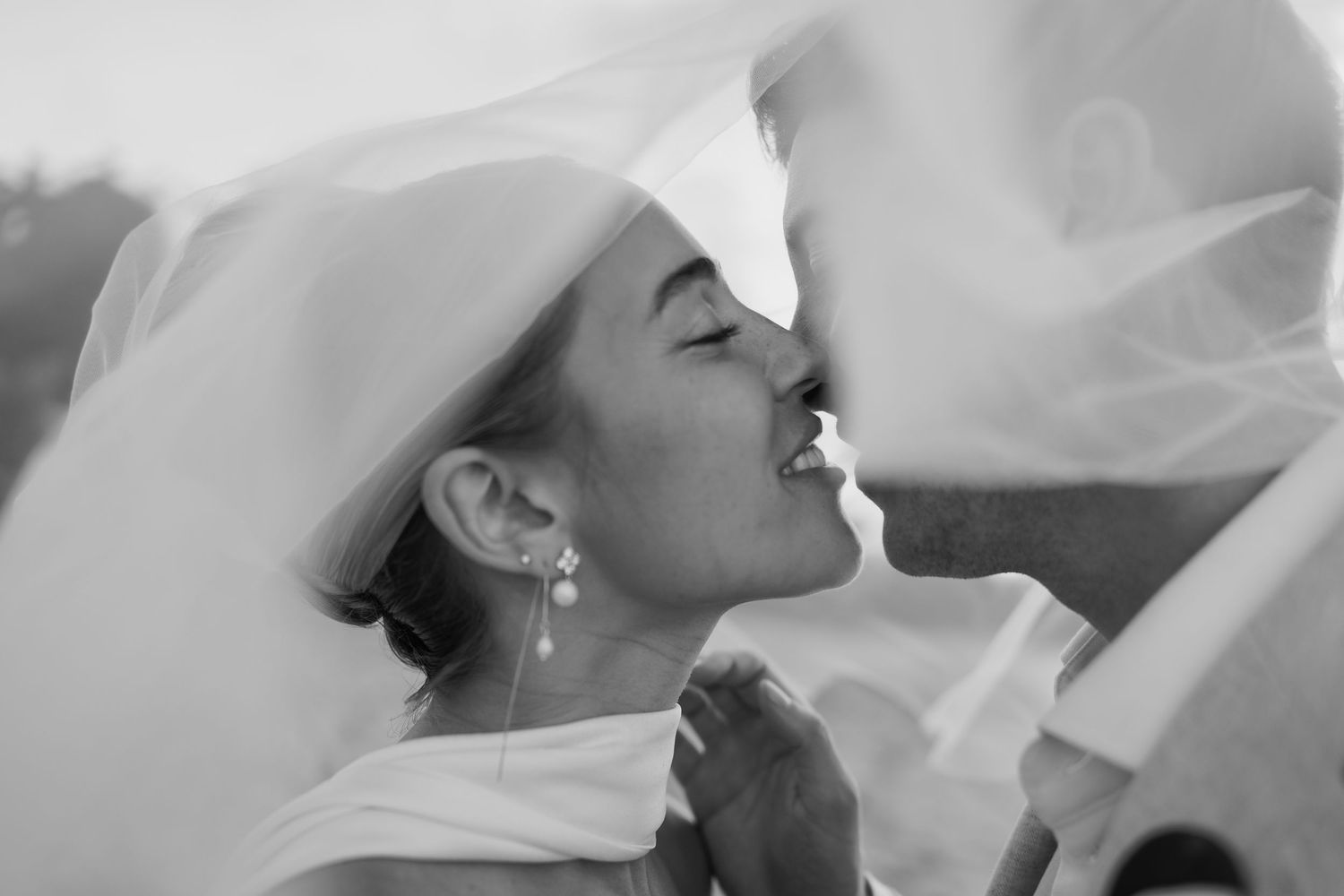 Black and white close-up of a bride kissing her partner beneath a flowing veil, showcasing elegant drop earrings.