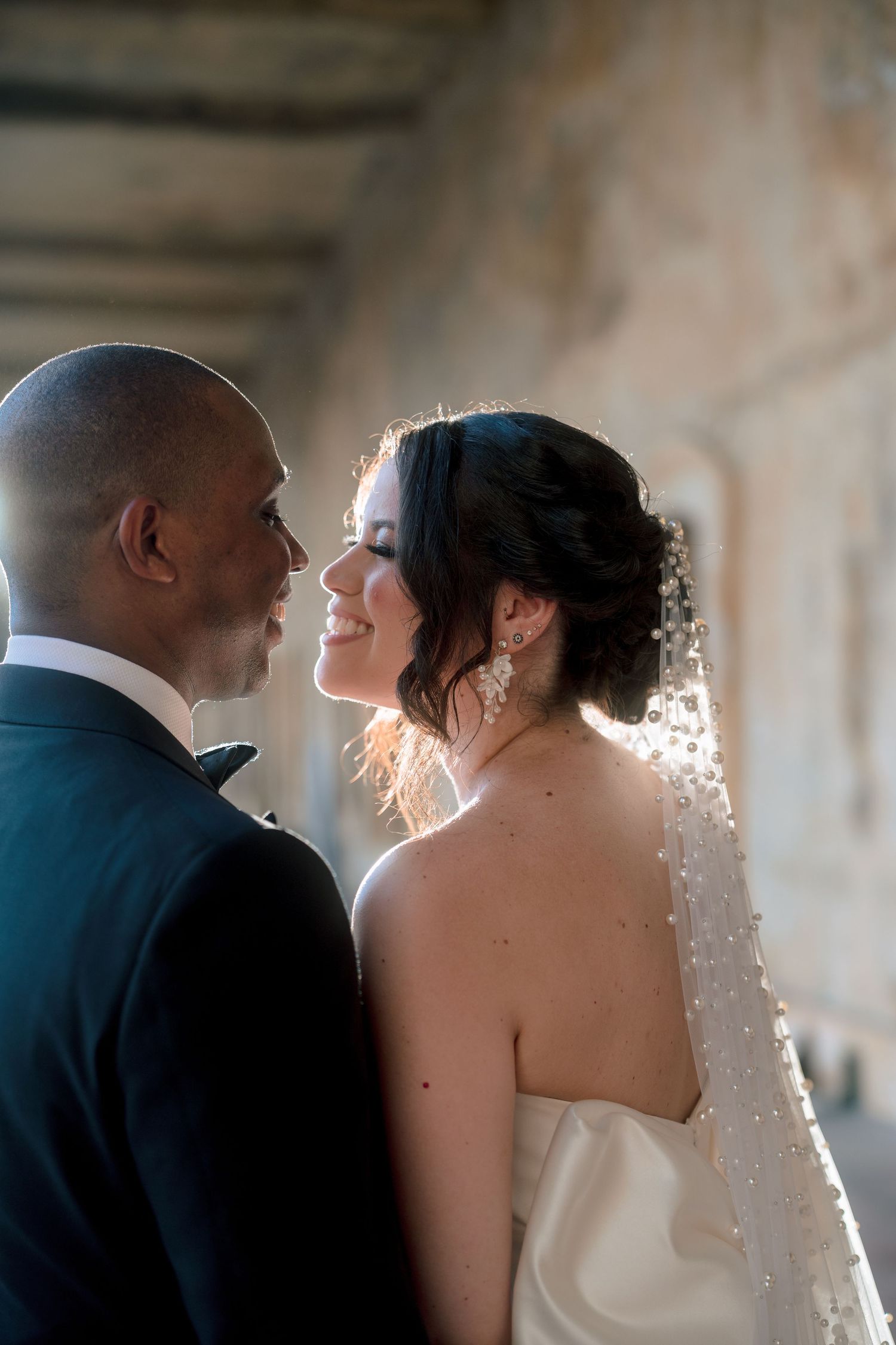 A bride and groom share an intimate moment, backlit by golden light, as she smiles up at him in a beaded veil.