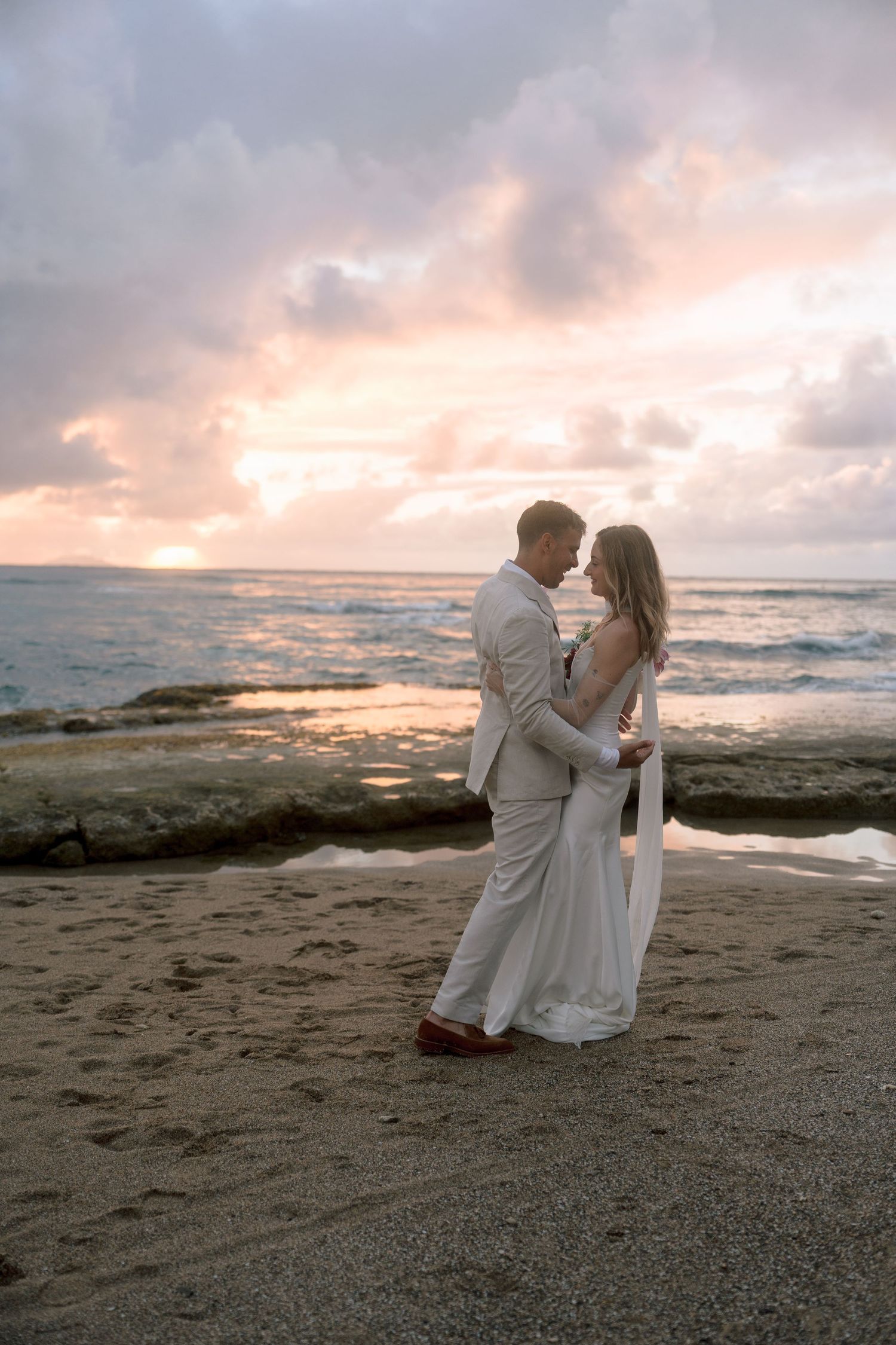 Newlyweds embracing on sandy beach at sunset with dramatic pink and purple cloudy sky backdrop.