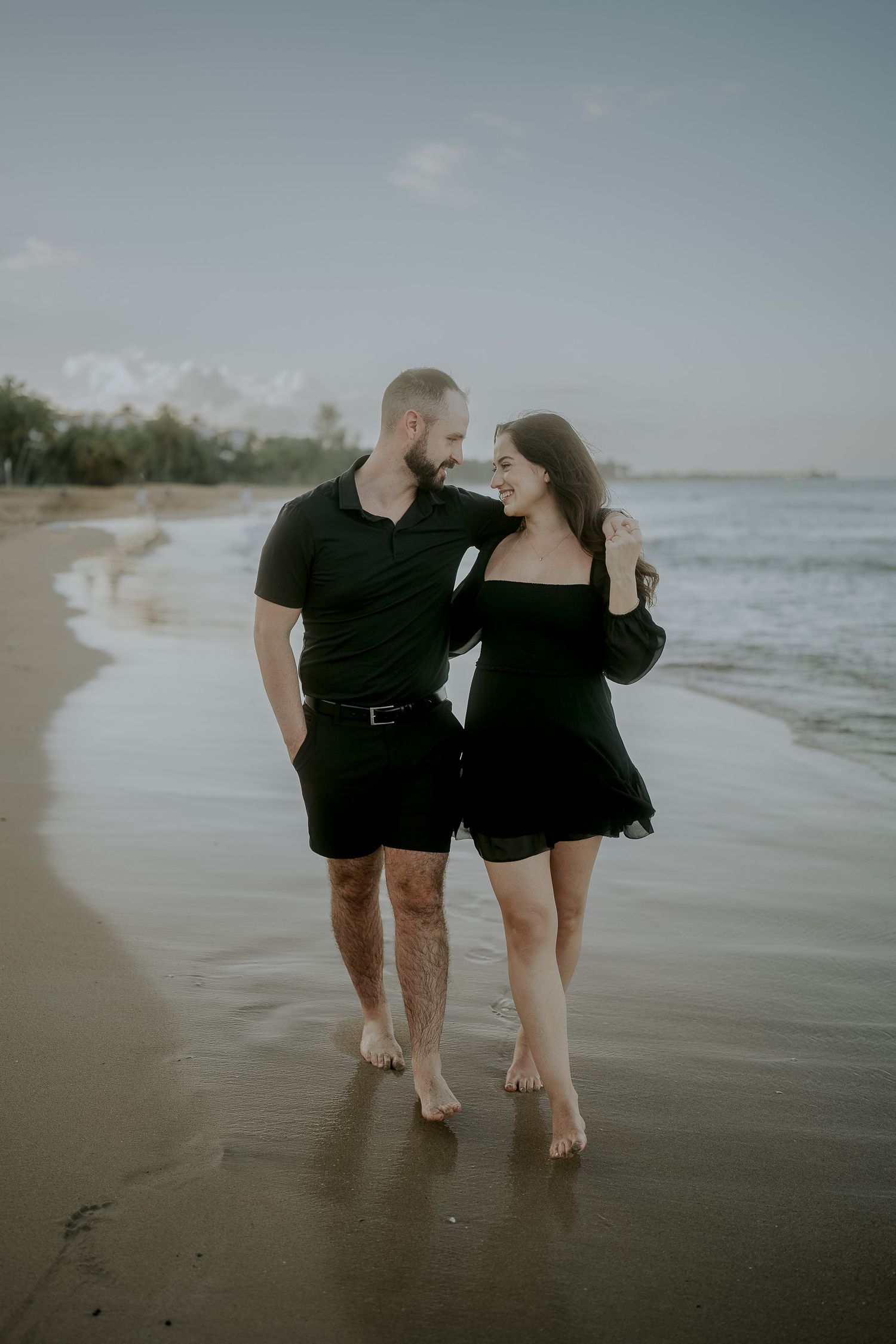 Couple in matching black outfits walking barefoot on a sandy beach shore, sharing a romantic moment at dusk.