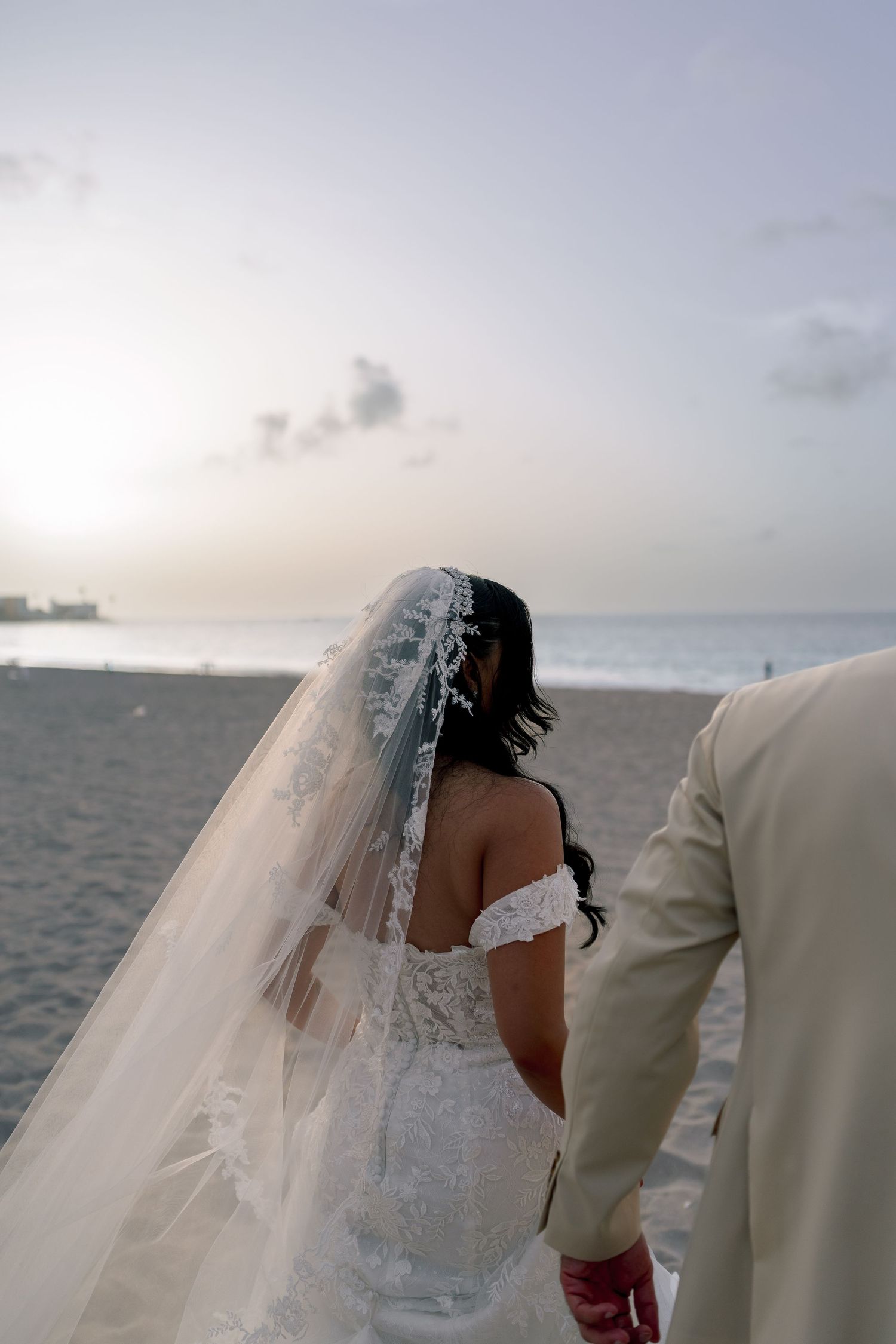 Bride in off-shoulder lace gown and cathedral veil walks with groom along sandy beach at sunset.