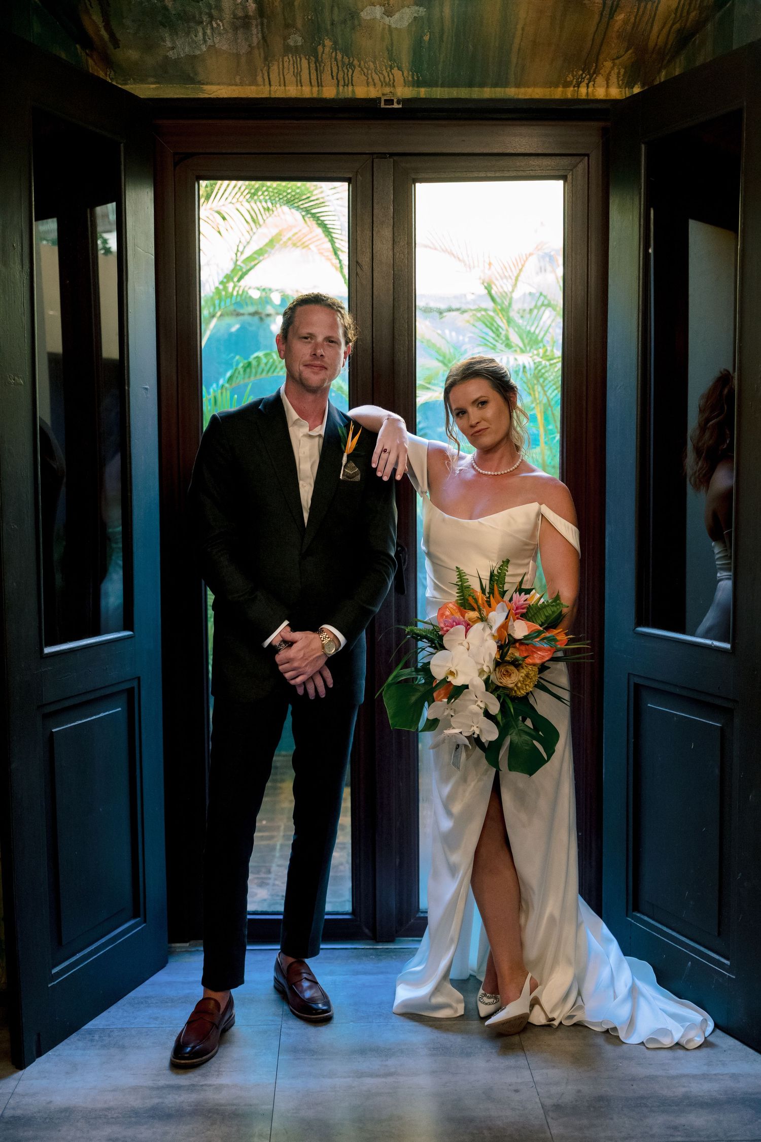 Bride and groom posing together in a doorway, bride holding a tropical floral bouquet.
