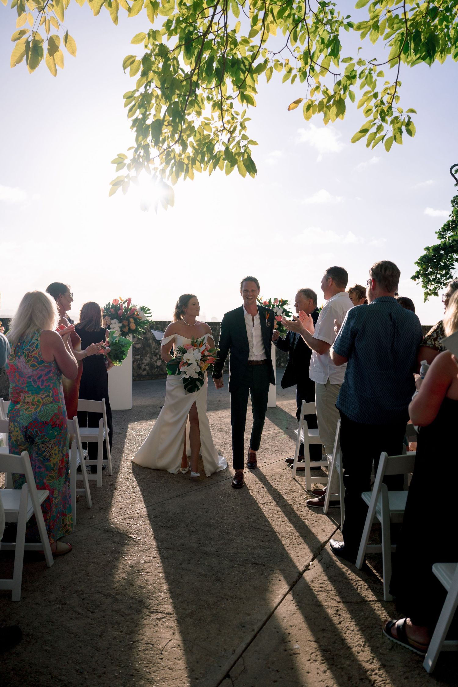 Newlyweds walk down outdoor aisle at sunset as guests celebrate their wedding ceremony.