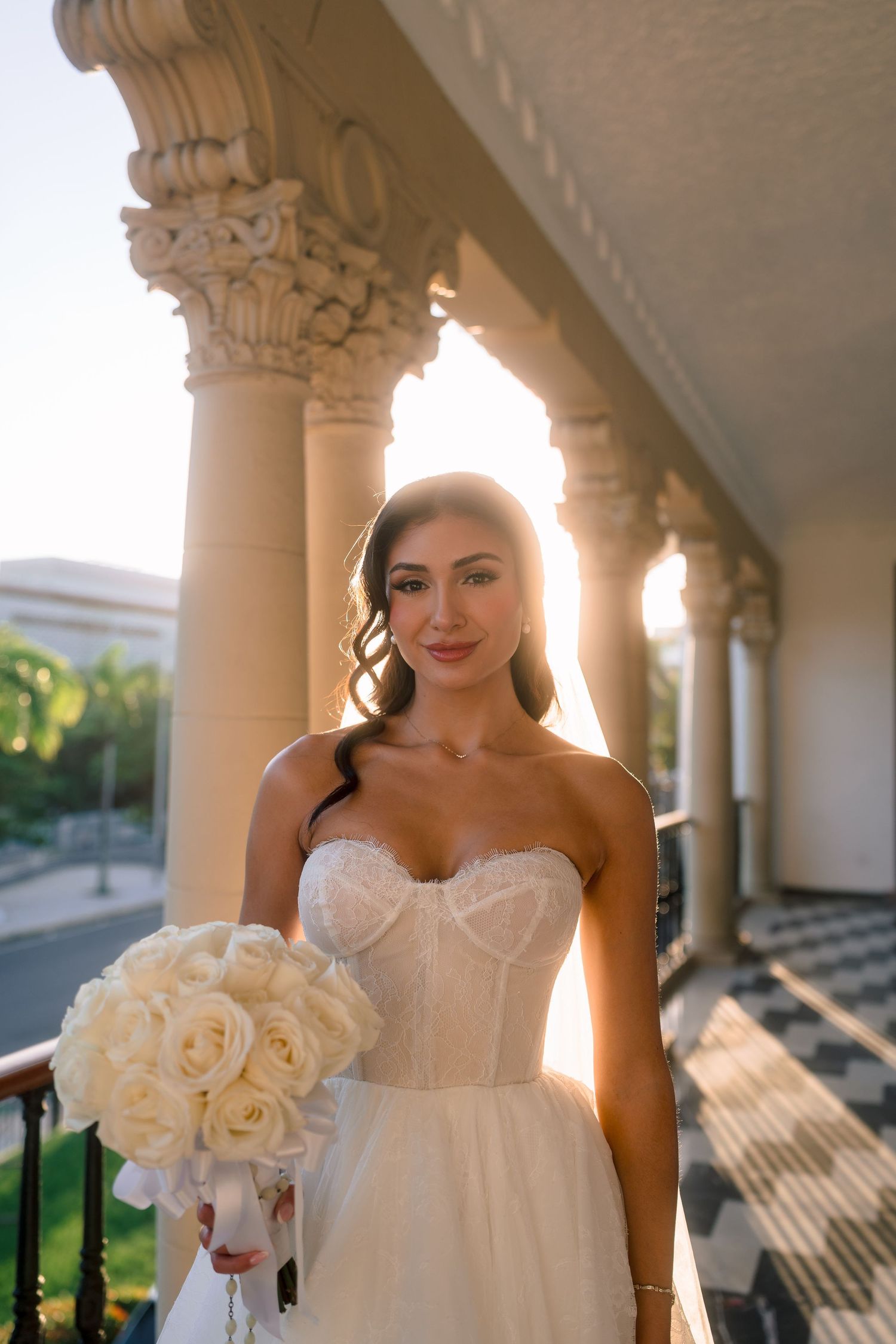 Bride in strapless lace corset gown holds white rose bouquet on sunlit classical colonnade.