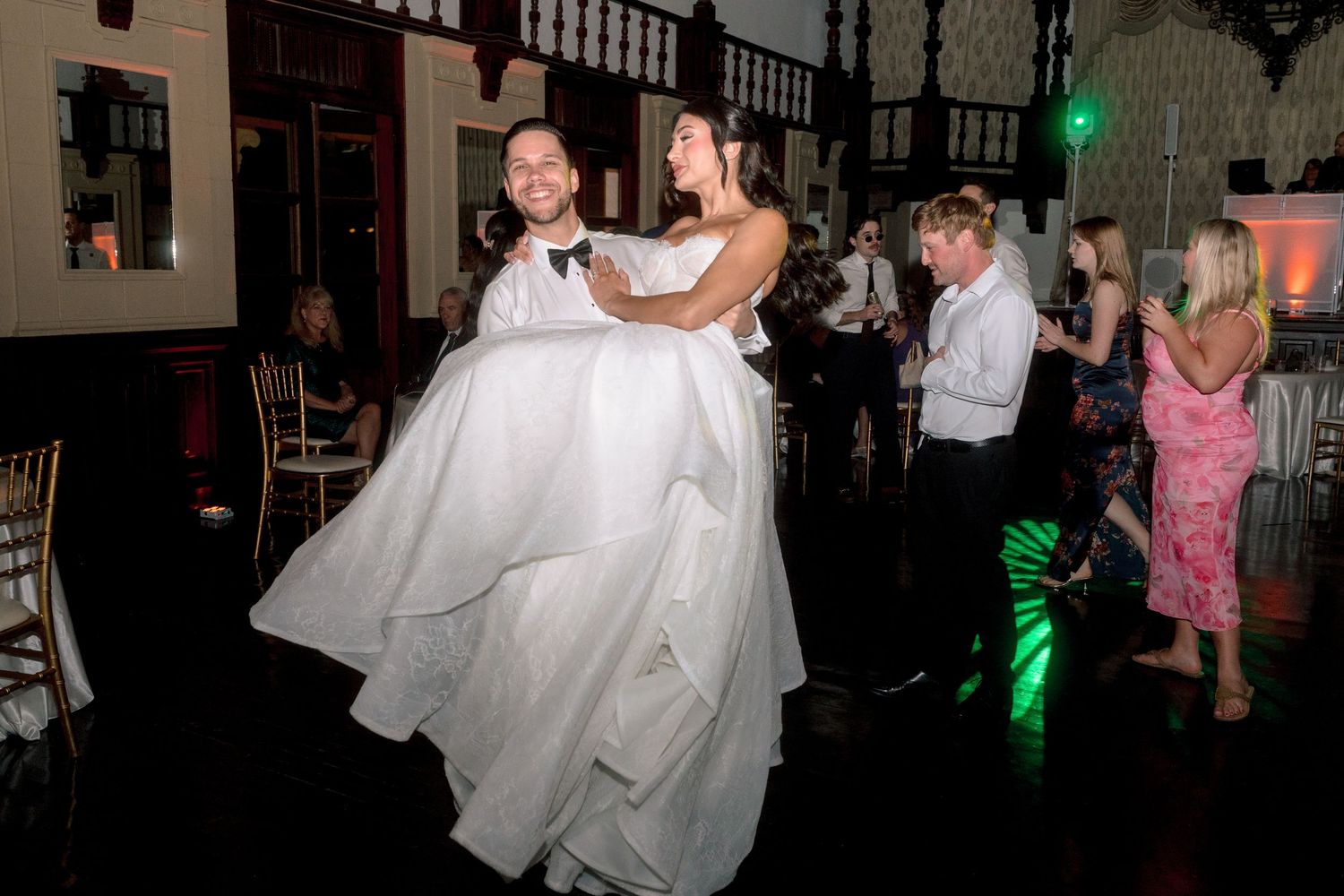 Groom lifts bride in white wedding gown on dance floor surrounded by celebrating guests.