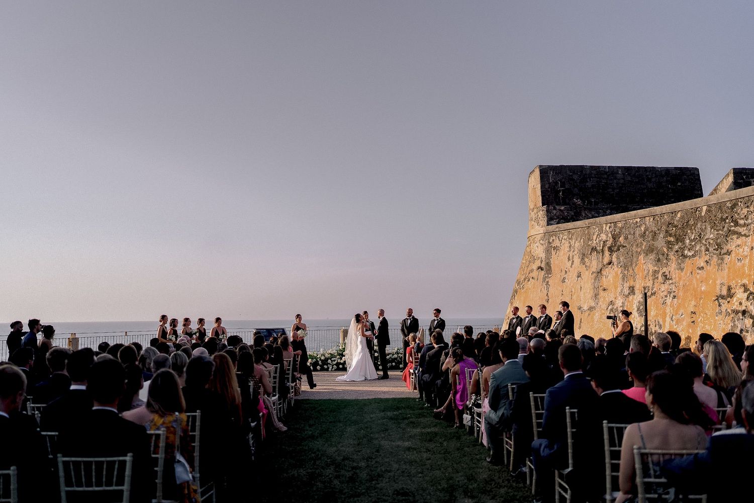 Outdoor wedding ceremony at sunset beside an ancient stone fort wall with ocean views and seated guests.