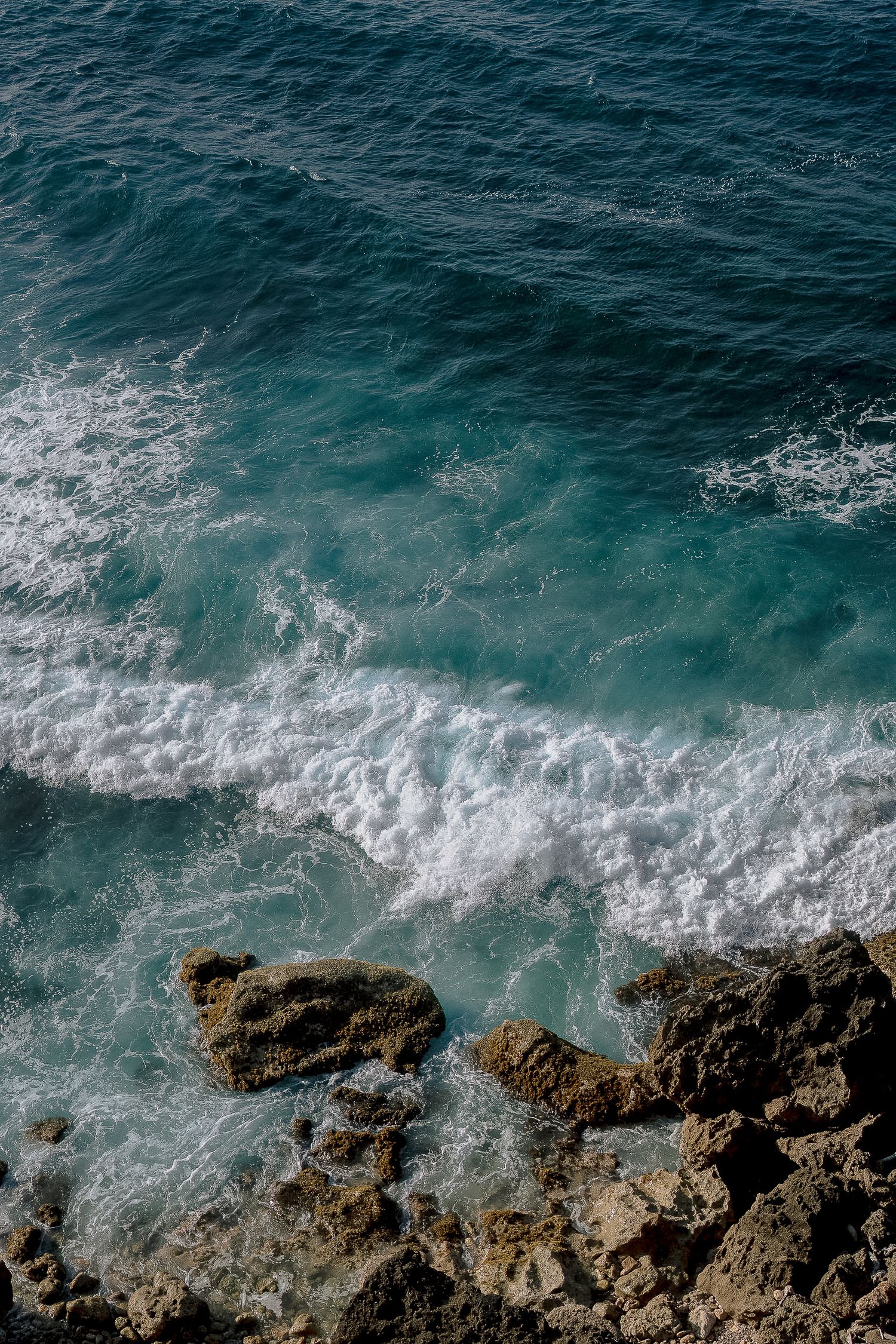 Turquoise ocean waves crashing against rocky shoreline from aerial view.