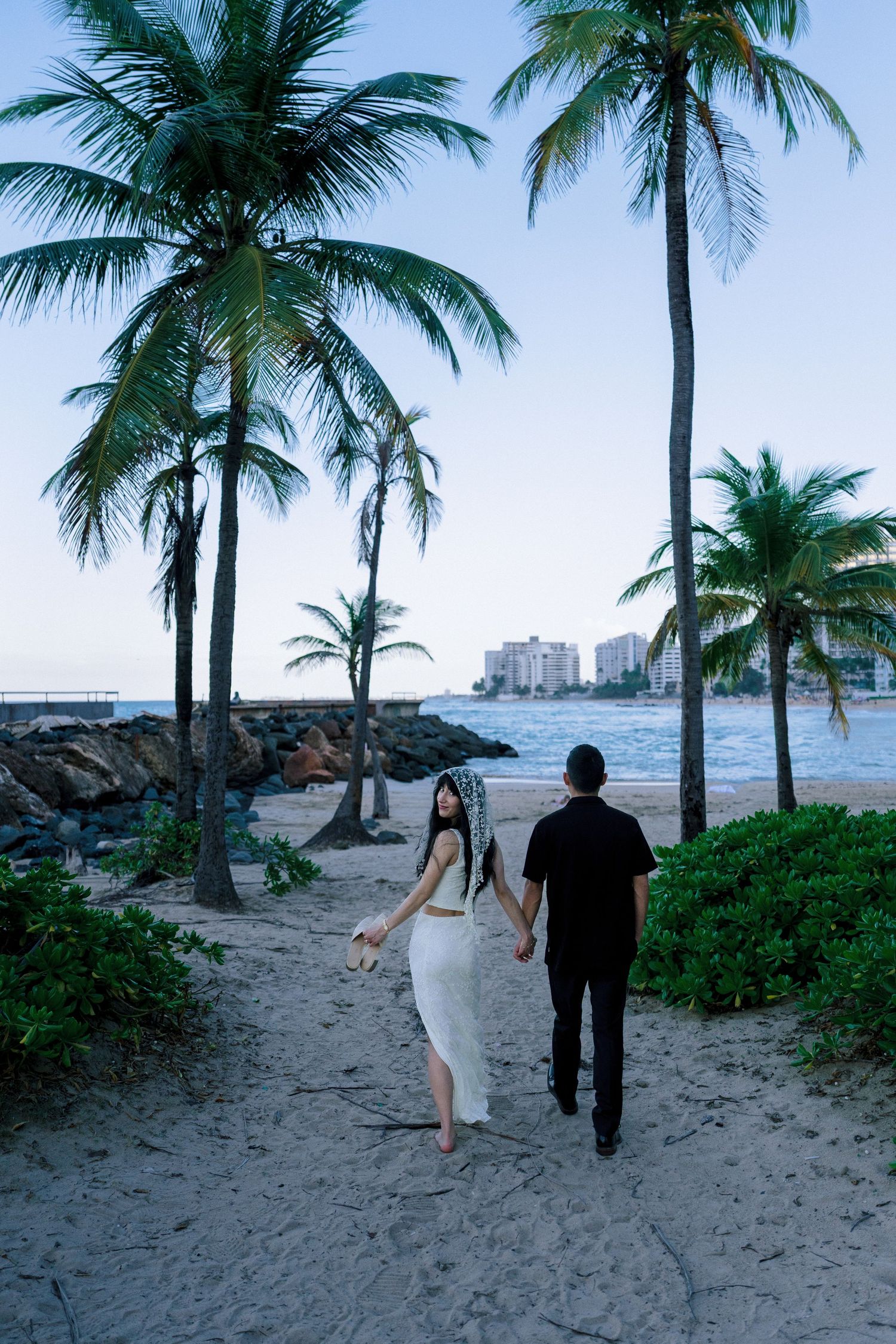 Couple in wedding attire holding hands on a tropical beach with palm trees and ocean views at dusk.