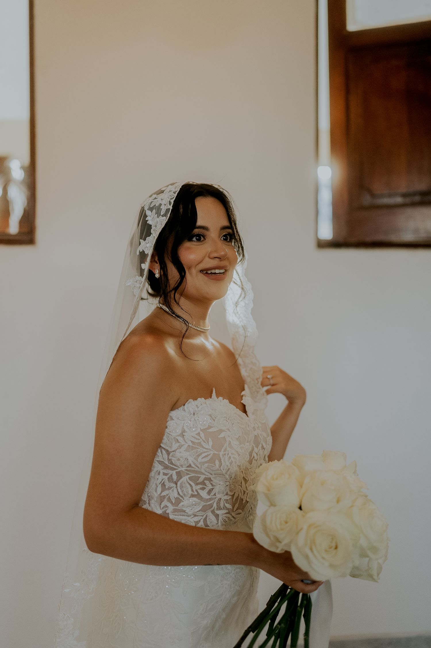 Smiling bride in lace strapless gown holding white rose bouquet wearing a delicate lace mantilla veil.