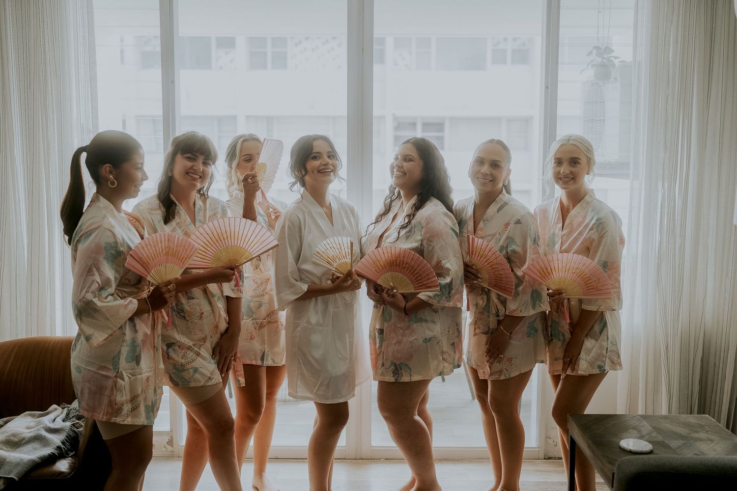 Bridesmaids in matching floral robes holding pink paper fans pose together in a bright room before a wedding ceremony.