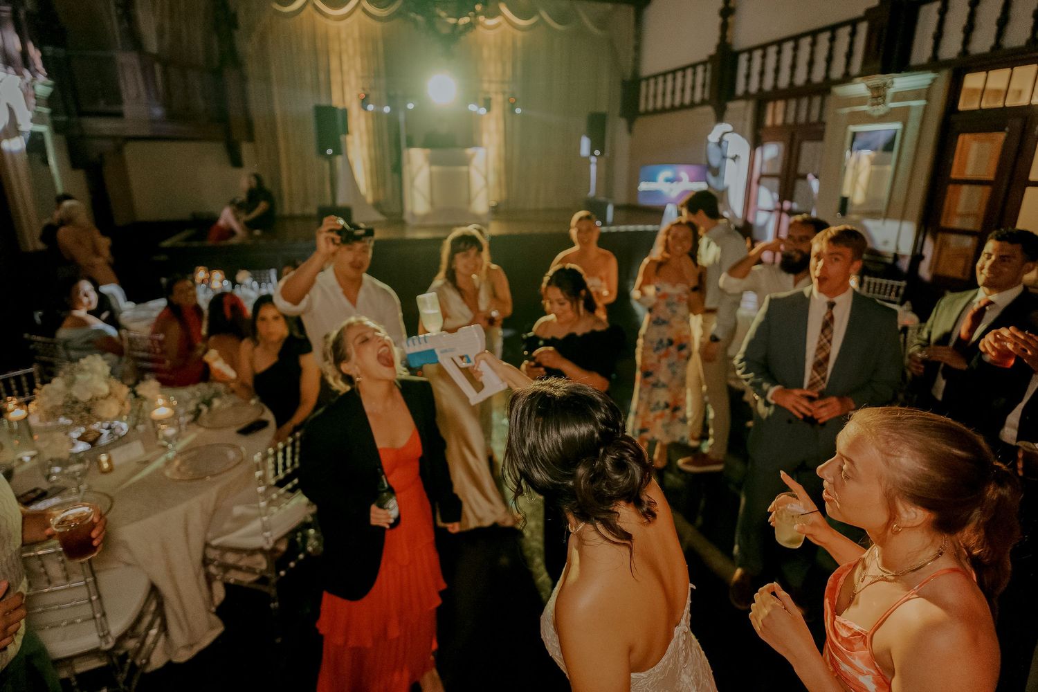 Wedding guests laugh and dance during lively reception party with colorful lighting and music.