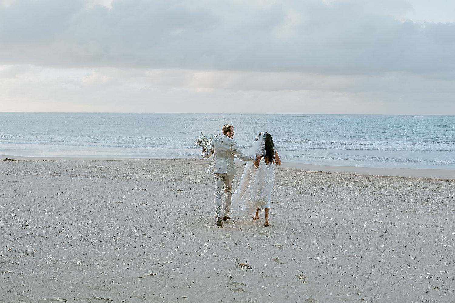 Bride and groom walking hand in hand on a sandy beach, with the ocean and cloudy sky in the background.