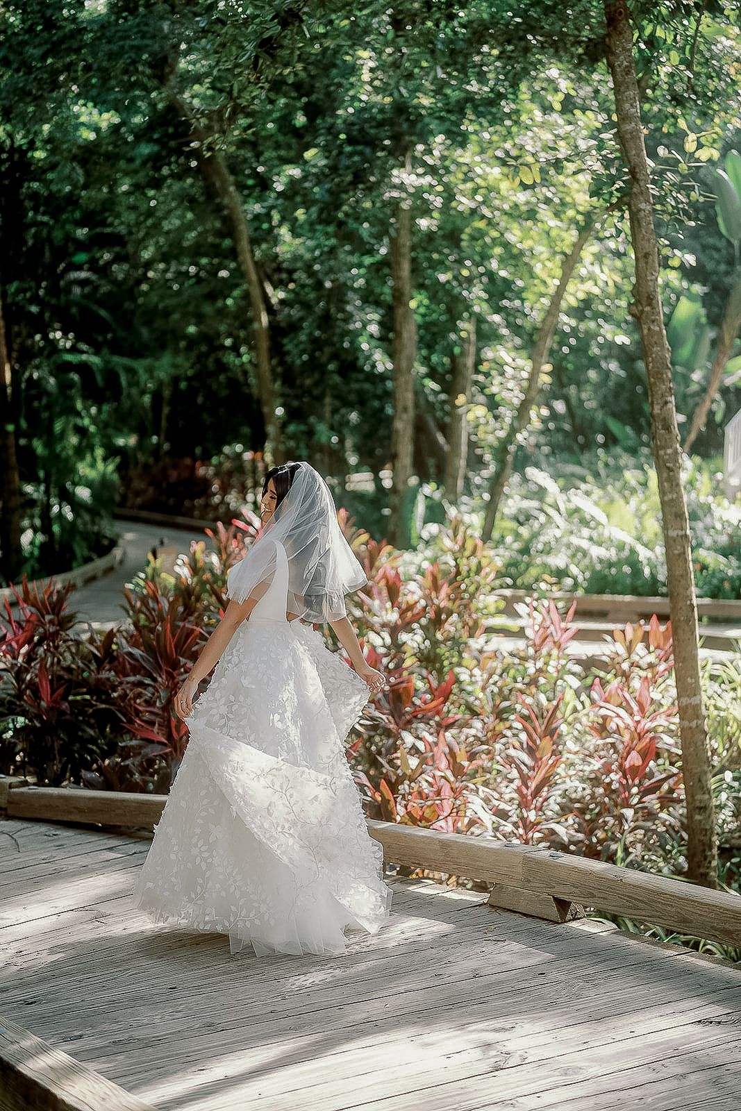 Bride in a white lace gown and veil standing on a wooden deck surrounded by lush tropical greenery.