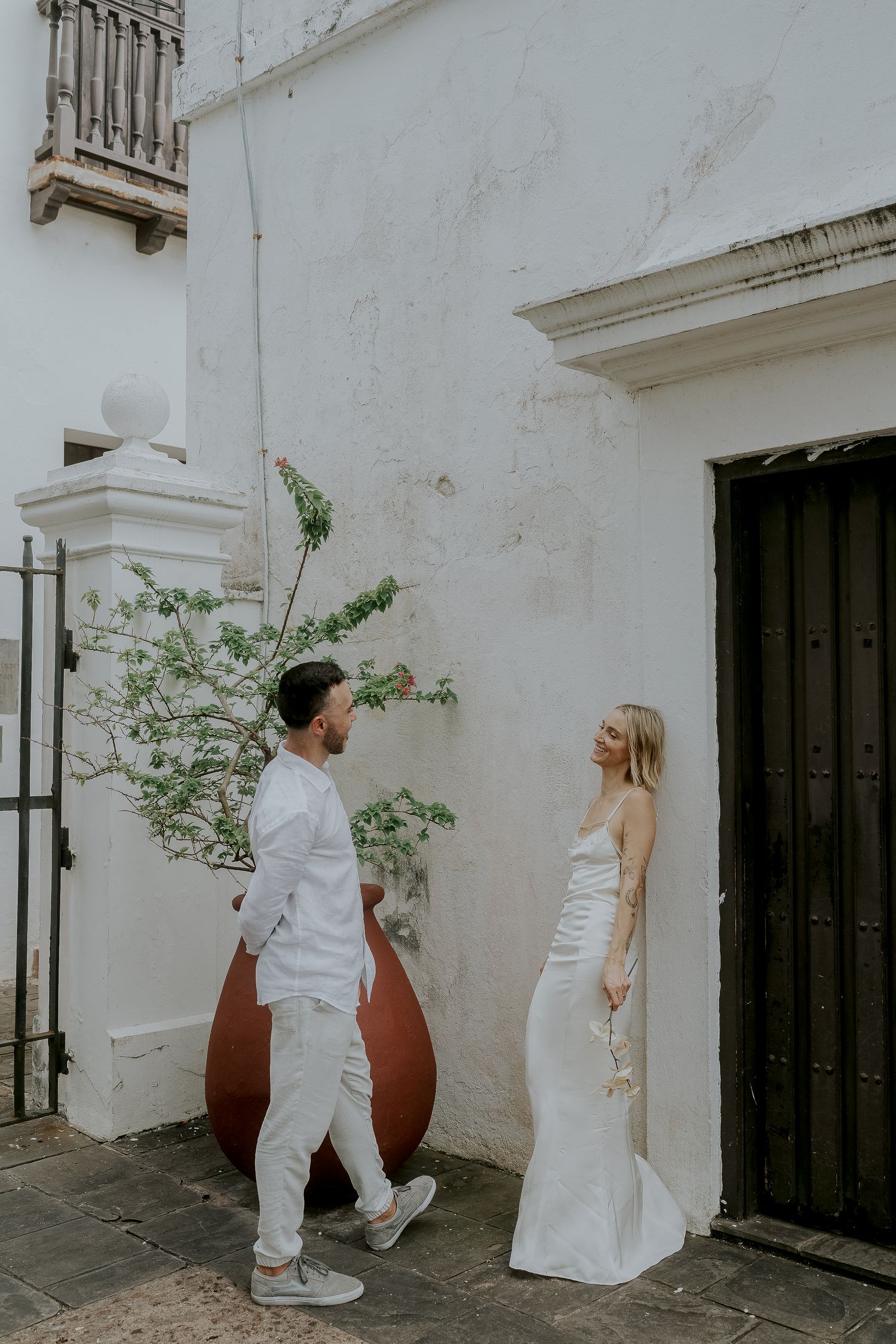 Bride in white satin gown and groom in white linen suit share a romantic moment by a whitewashed wall.