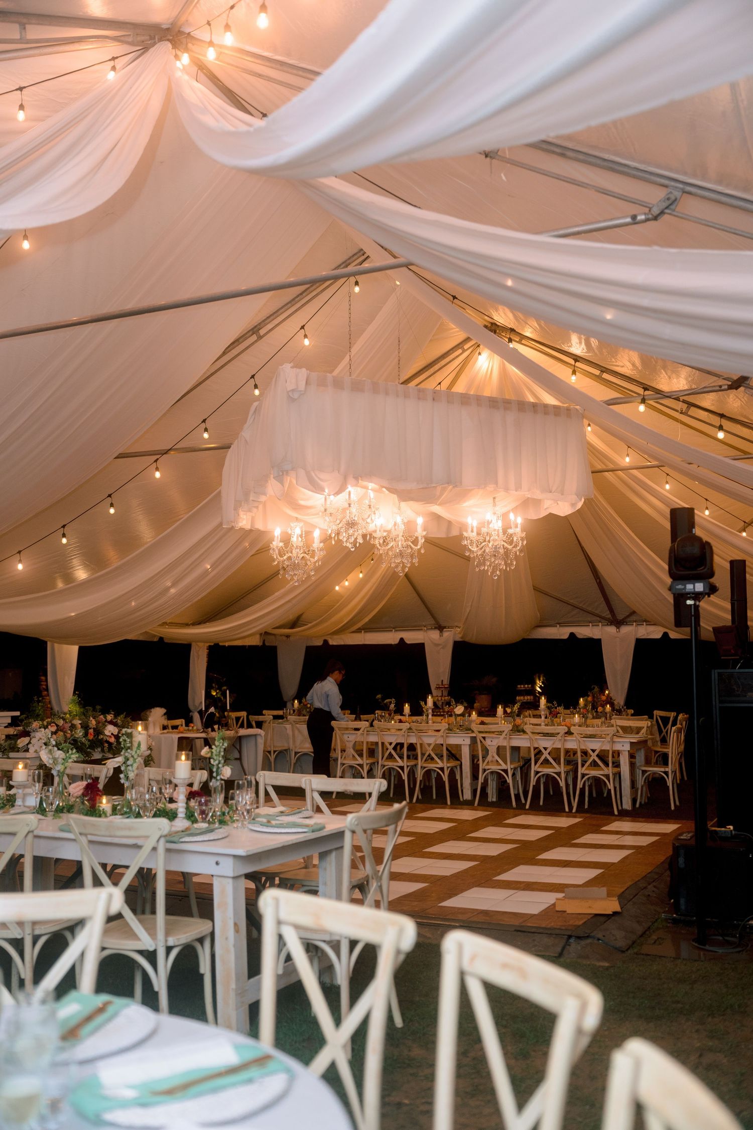 Elegant wedding reception tent with white draping, chandeliers, string lights, and white cross-back chairs.