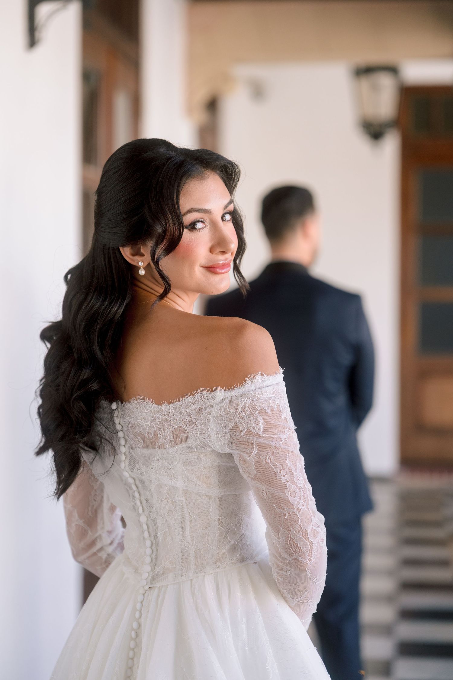 Bride in off-shoulder lace wedding dress looks back smiling, groom in navy suit stands blurred in background.