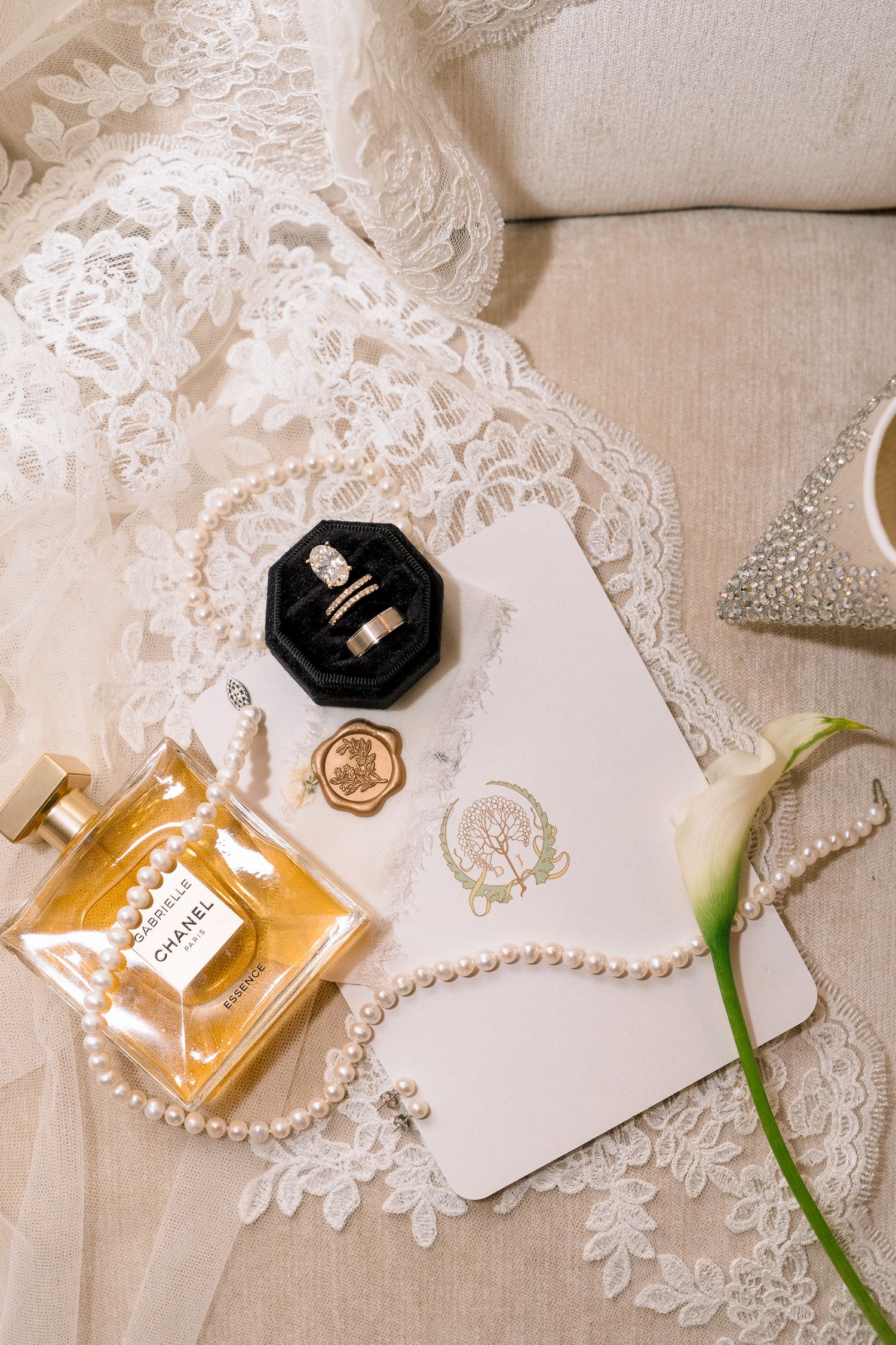 Elegant bridal flat lay with Chanel perfume, pearl necklace, wedding rings, wax seal, invitation, calla lily, and lace gown.