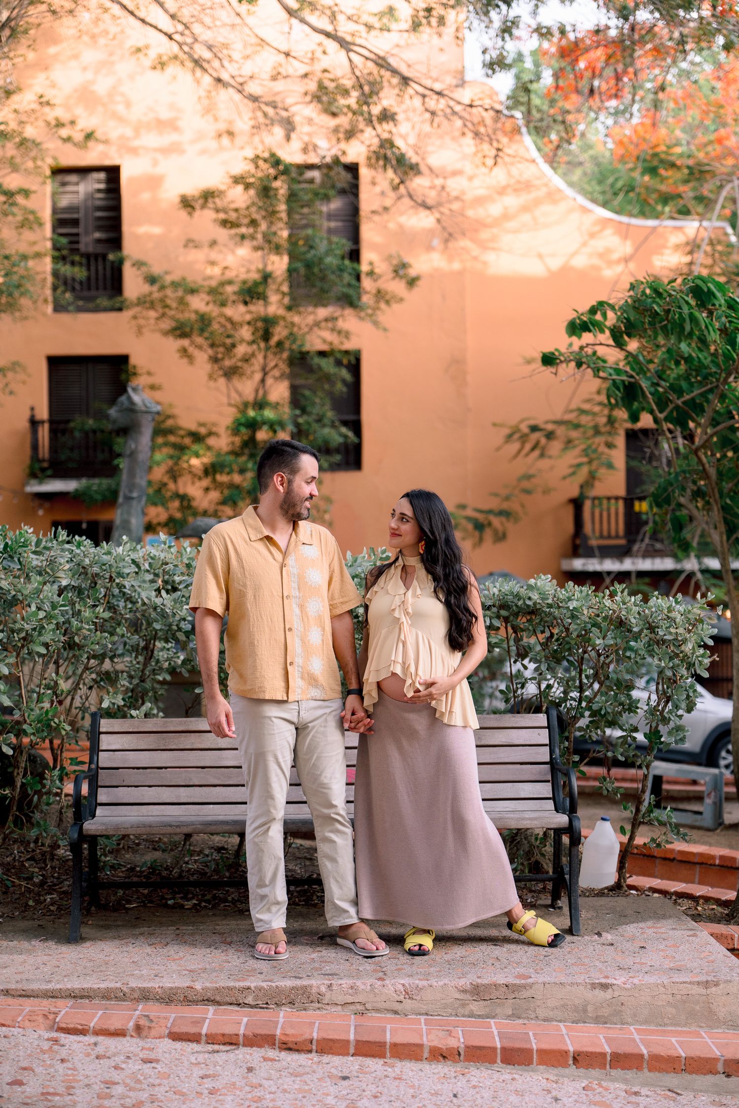 Couple holding hands smiling at each other near a bench outside an orange building surrounded by greenery.