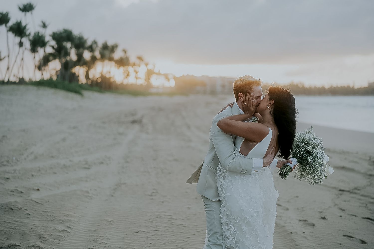 Newlywed couple sharing a romantic kiss on a sandy beach at sunset with tropical palm trees in the background.