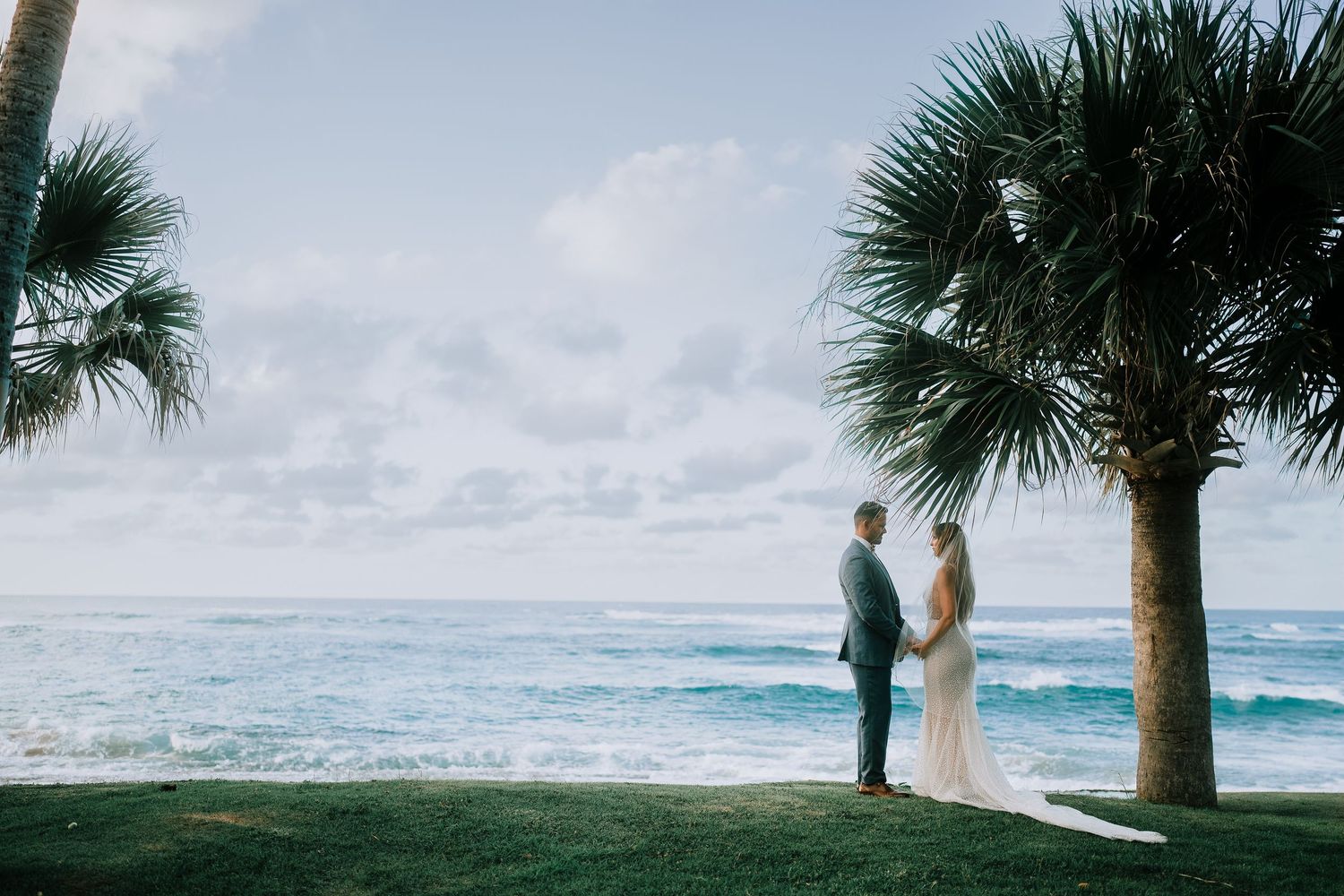 Bride and groom standing beneath palm tree overlooking stunning ocean waves on wedding day.
