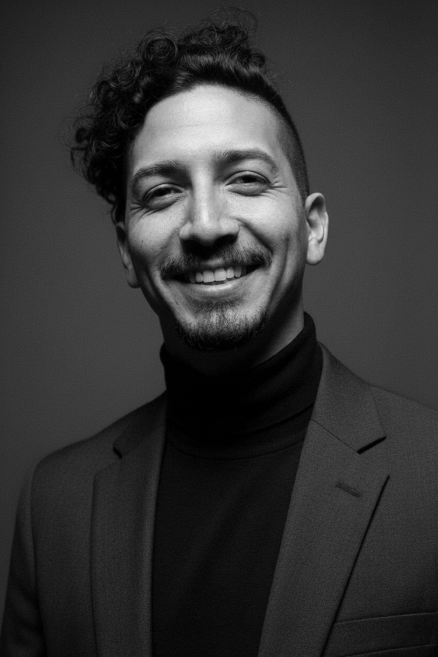 Black and white portrait of a smiling man with curly hair wearing a turtleneck and blazer against a dark background.