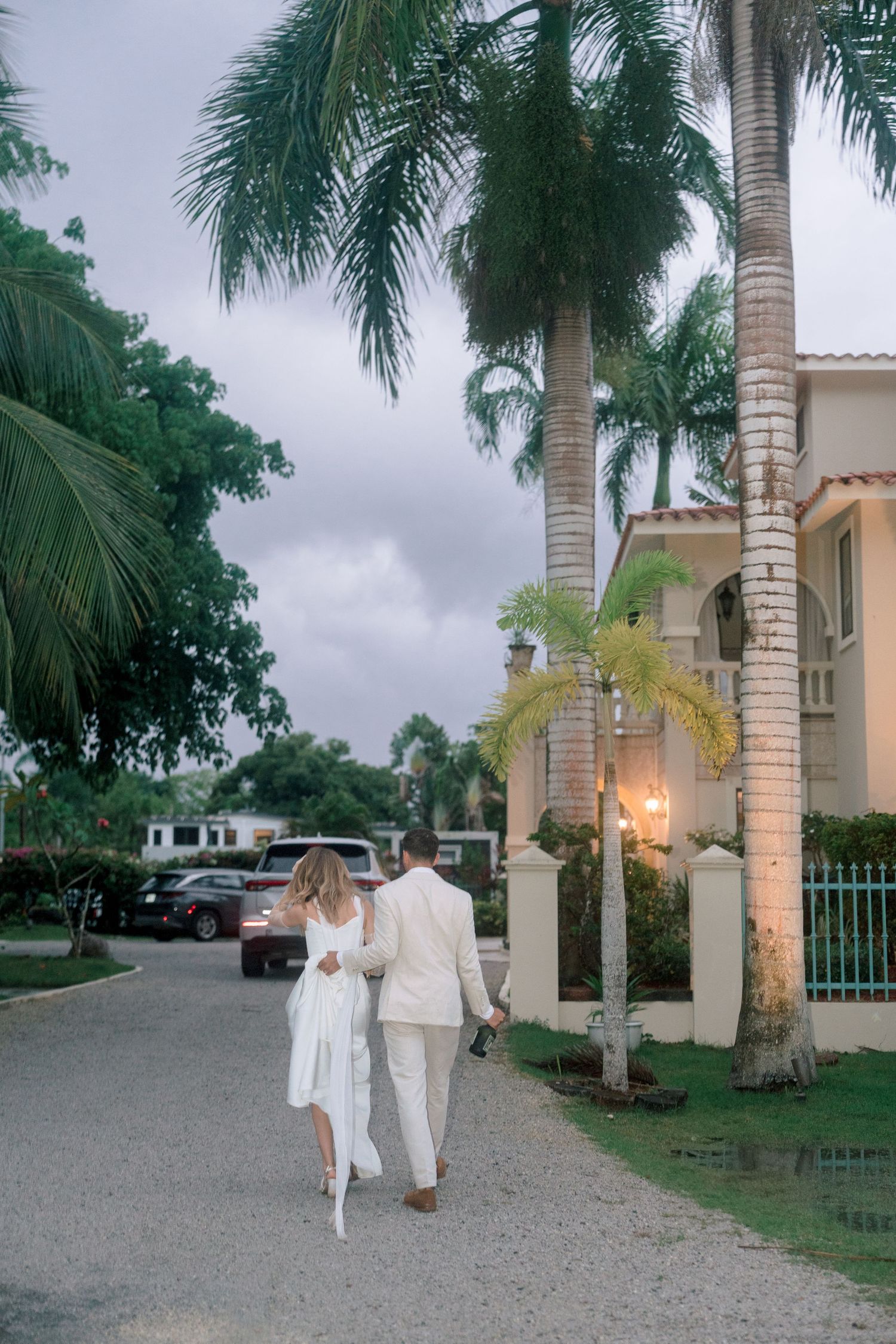 Newlyweds walk hand in hand along palm tree-lined driveway of tropical wedding venue at dusk.