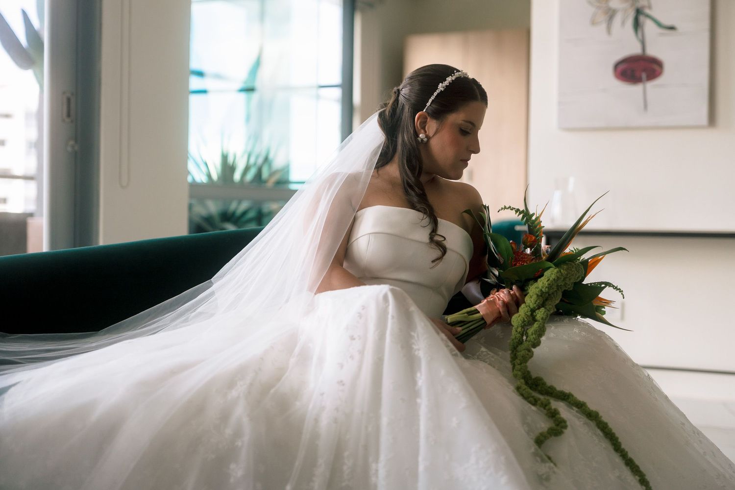 Bride in white strapless ball gown with veil holding colorful bouquet, seated elegantly in a softly lit room.