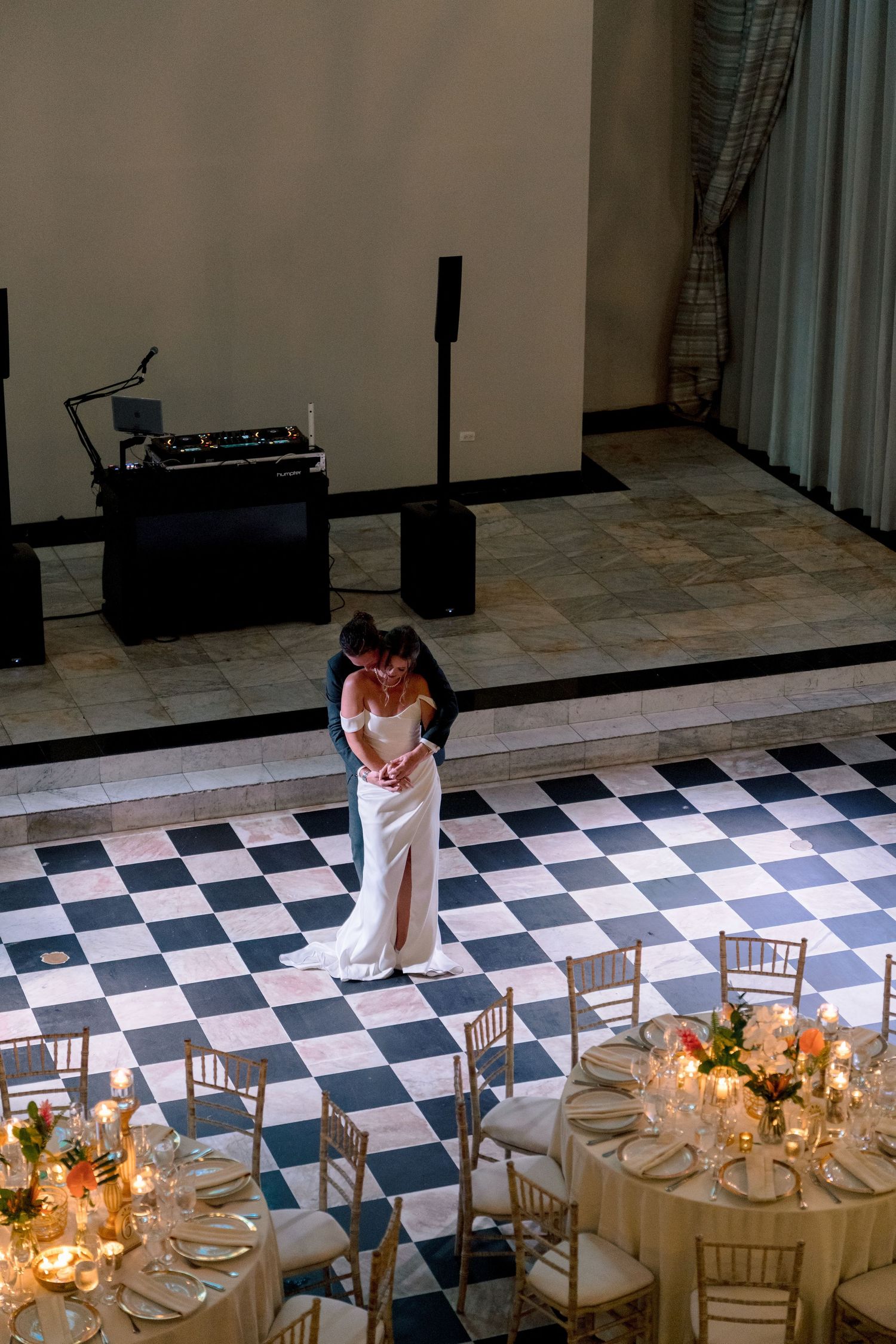 Bride and groom sharing first dance on black and white checkered floor surrounded by candlelit tables.