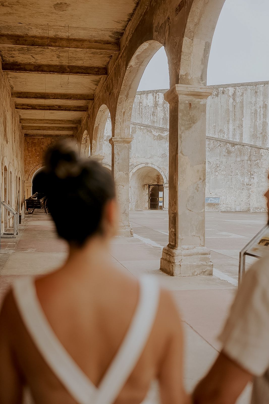 Bride in backless white dress walks through stone arched colonnade at historic venue.