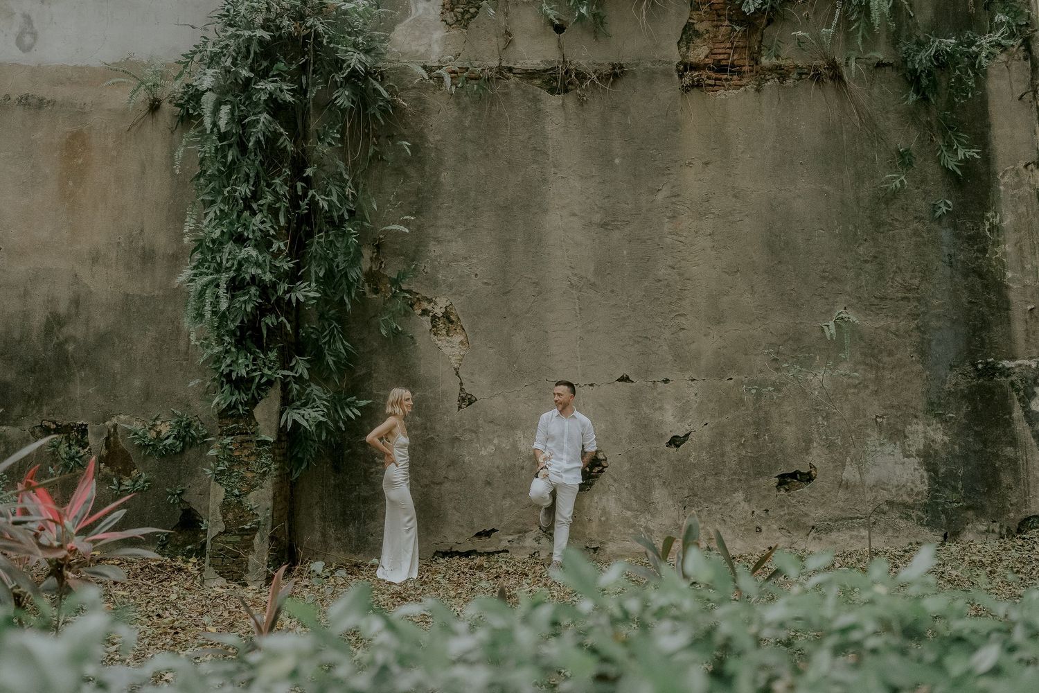 Couple in wedding attire standing among tropical plants against a weathered concrete wall in a lush garden setting.
