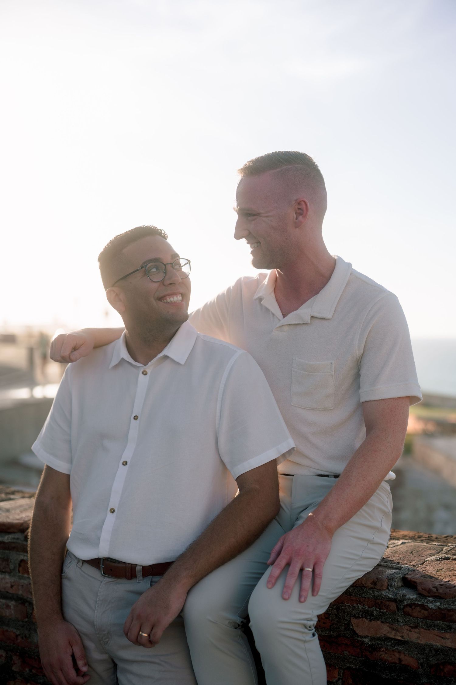 Two men in white shirts smile at each other while sitting outdoors in warm sunlight near a coastal backdrop.